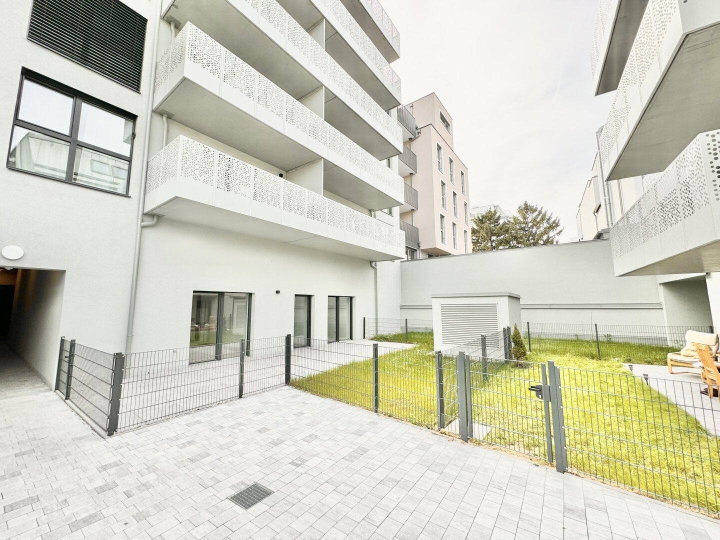 Außenbereich mit gepflasterter Terrasse, Grünfläche und moderner Wohnanlage im Hintergrund.
