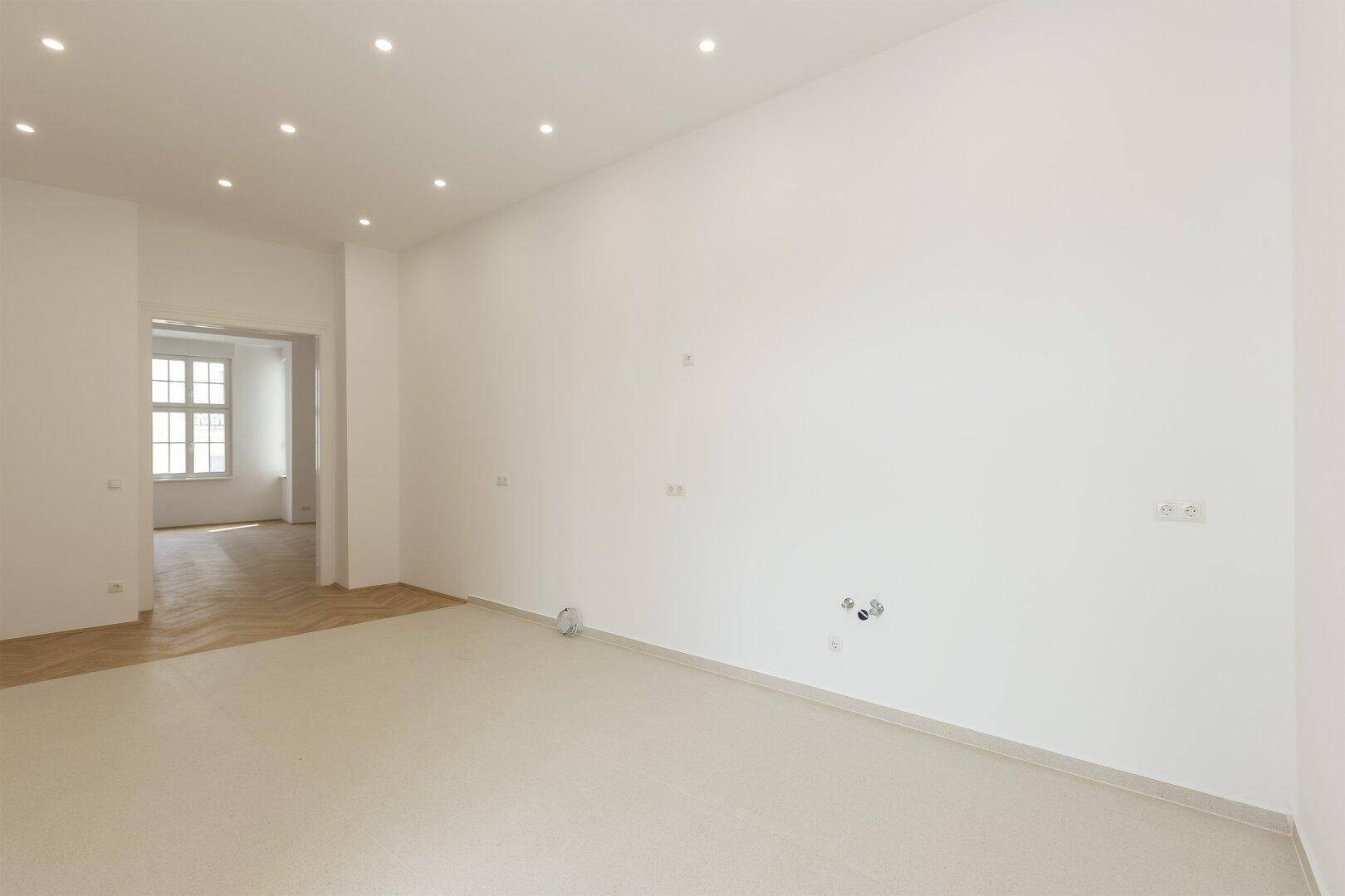 Helles, leeres Zimmer mit weißen Wänden, Einbaustrahlern und hellem Fliesenboden, angrenzend an einen Raum mit Parkett.