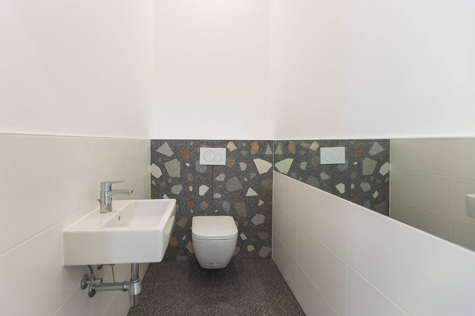 Gäste-WC mit wandhängender Toilette, kleinem Waschbecken und dekorativen Terrazzo-Fliesen an der Wand.