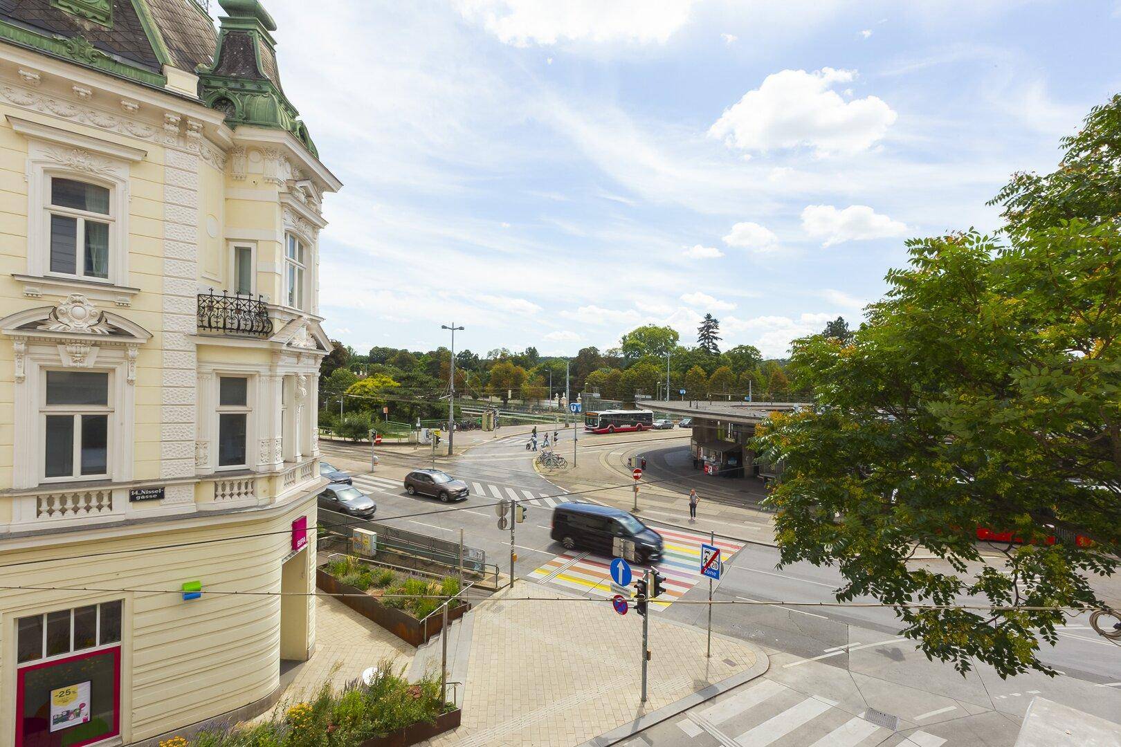 Blick von oben auf eine belebte Straßenkreuzung mit Verkehr, einem historischen Gebäude und Bäumen.