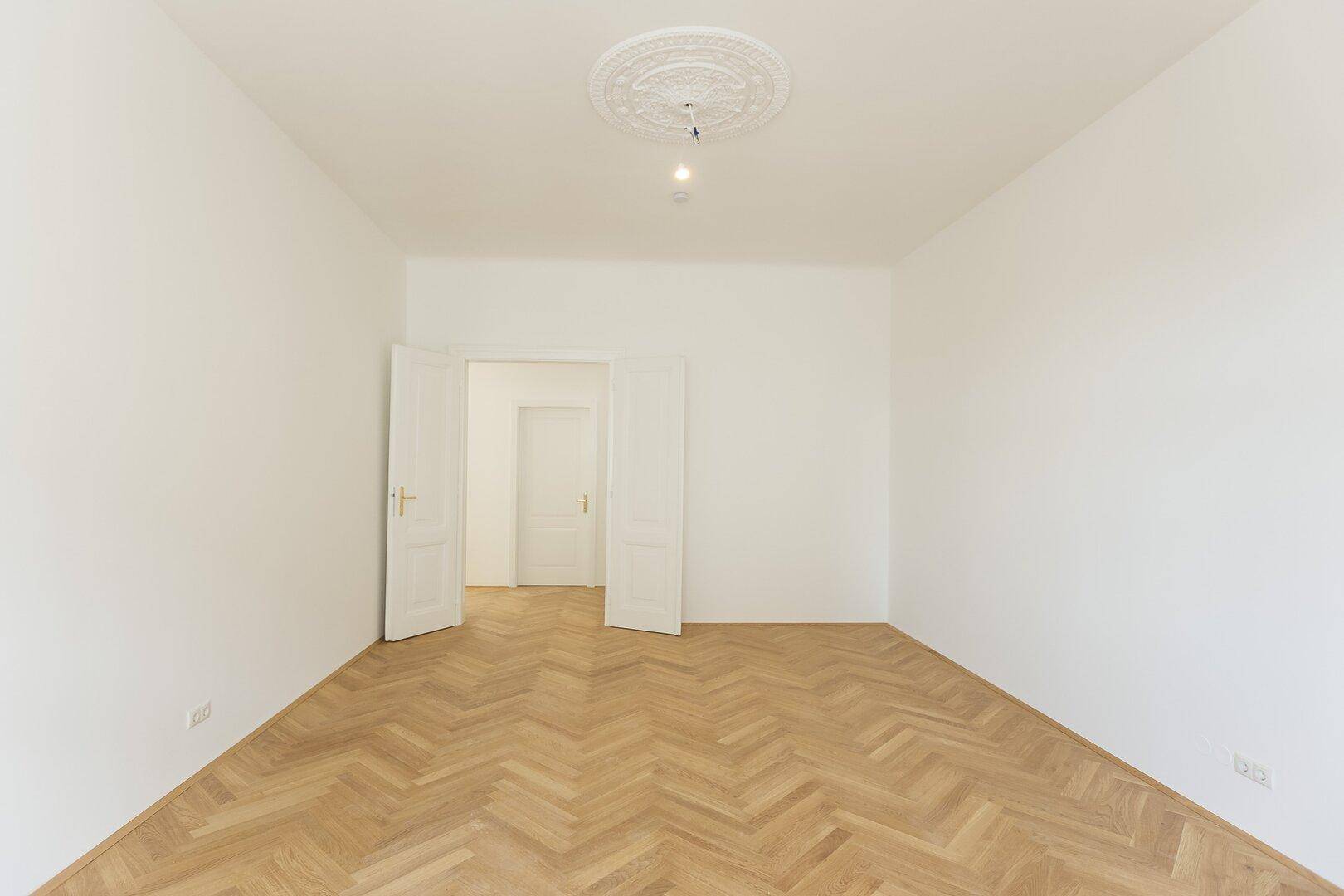 Helles Zimmer mit Fischgrätparkettboden, weißen Wänden und eleganten Doppeltüren, die zu einem weiteren Raum führen.