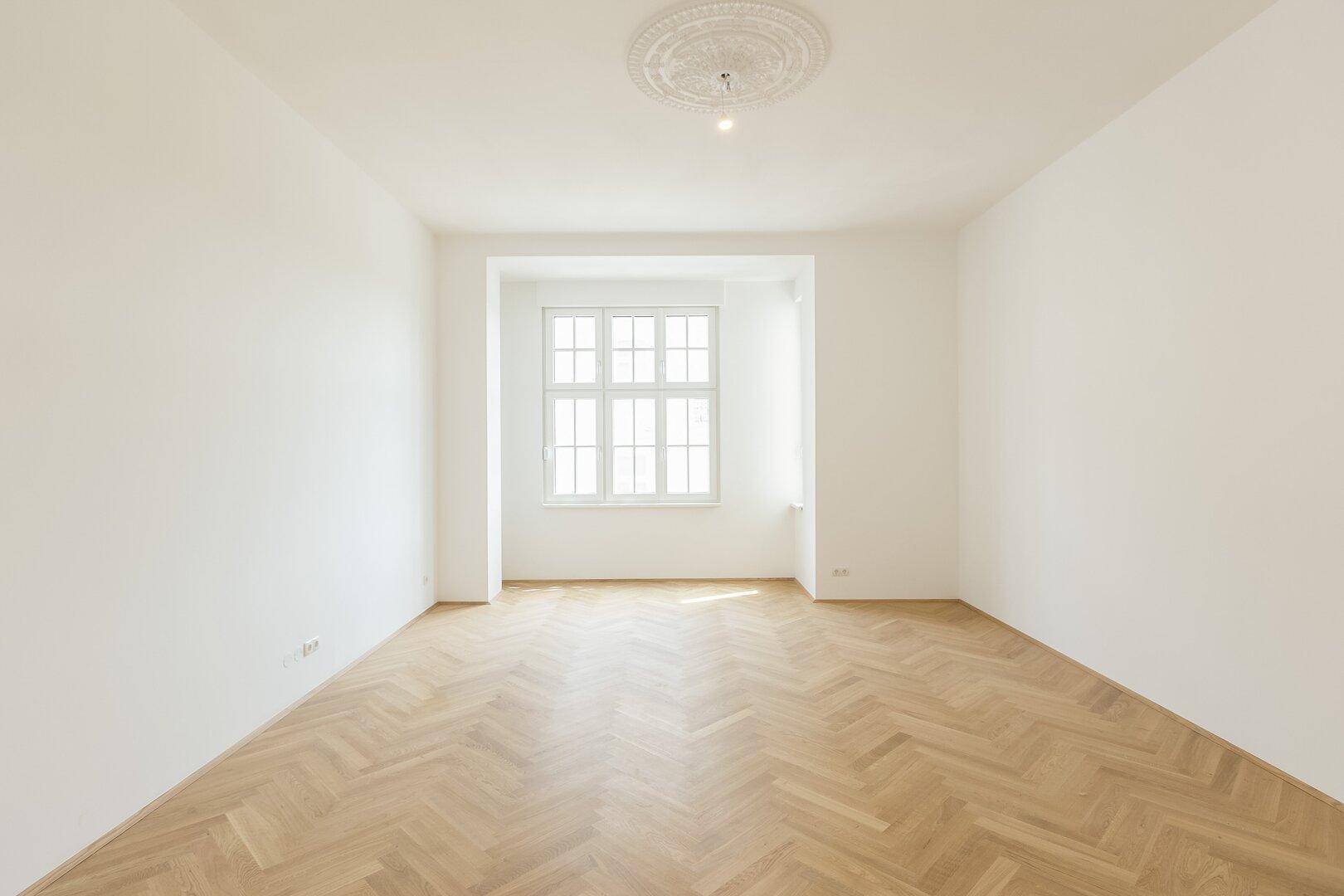 Geräumiges, helles Zimmer mit Fischgrätparkett, weiß gestrichenen Wänden und einem großen Fenster in einer Nische.