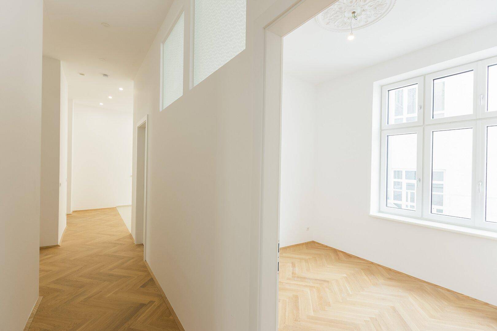 Heller Flur mit Einbaustrahlern und Blick in ein angrenzendes Zimmer mit Fischgrätparkett und Fenster.