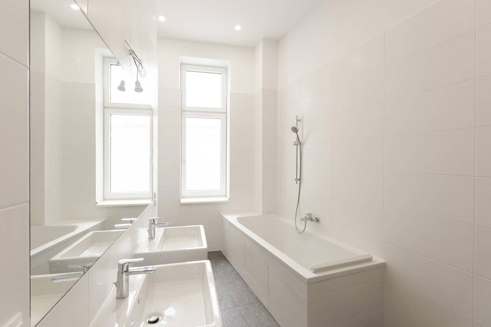 Modernes Badezimmer mit zwei Waschbecken, einer Badewanne, weißen Fliesen und zwei Fenstern für Tageslicht.