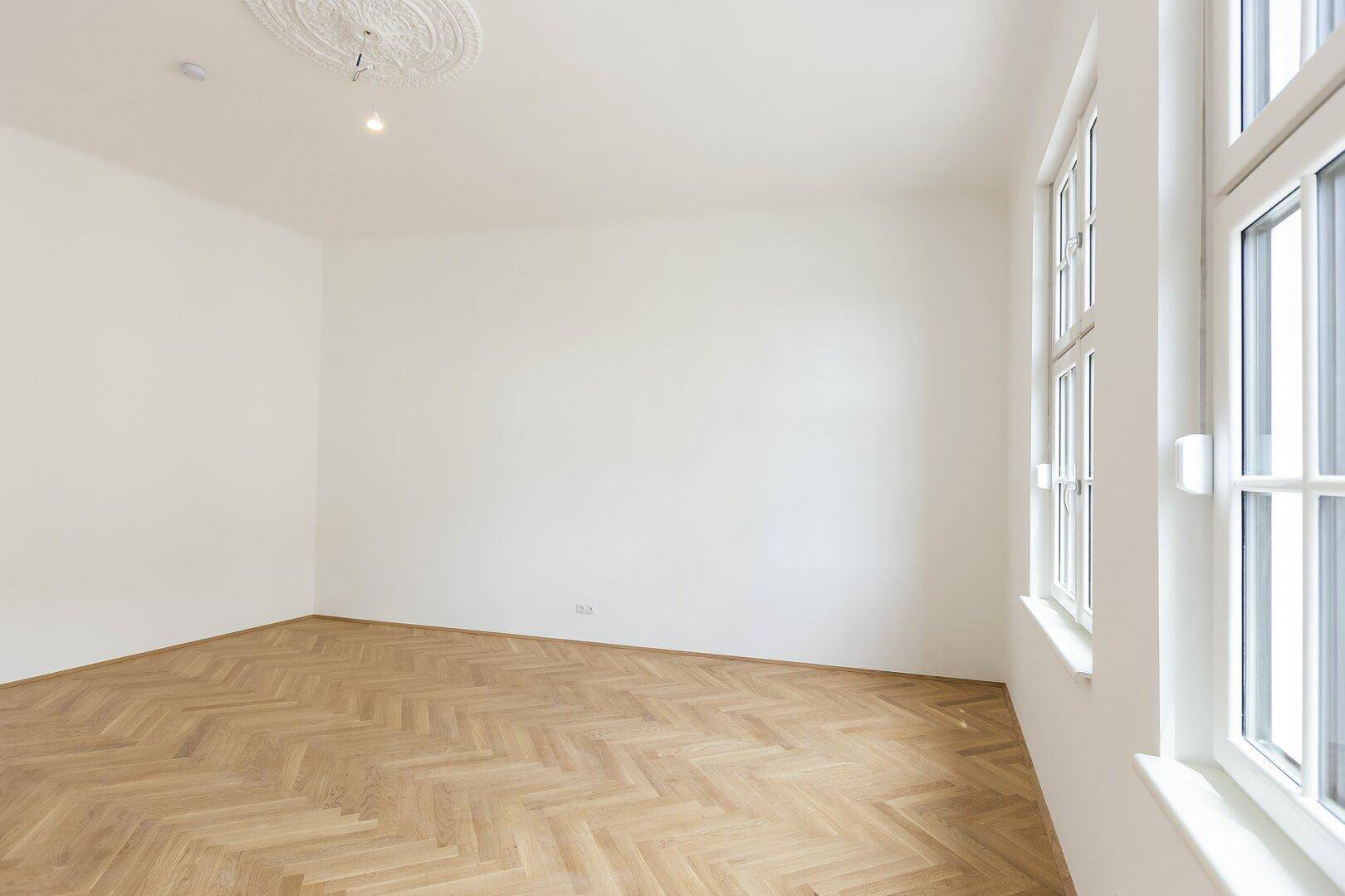 Geräumiges, helles Zimmer mit Fischgrätparkettboden, weißen Wänden und einem großen Fenster, das viel Licht hereinlässt.
