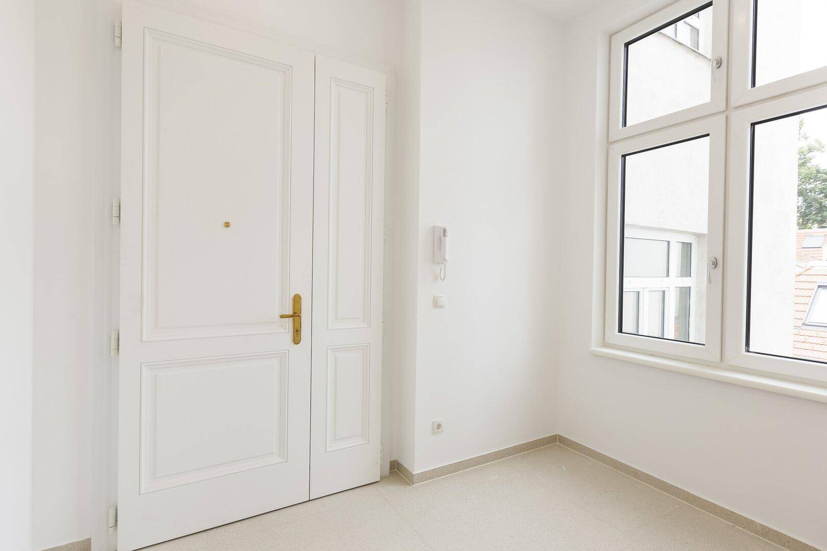 Eingangsbereich mit weißer Doppeltür, Gegensprechanlage und einem Fenster, der mit hellen Fliesen ausgelegt ist.