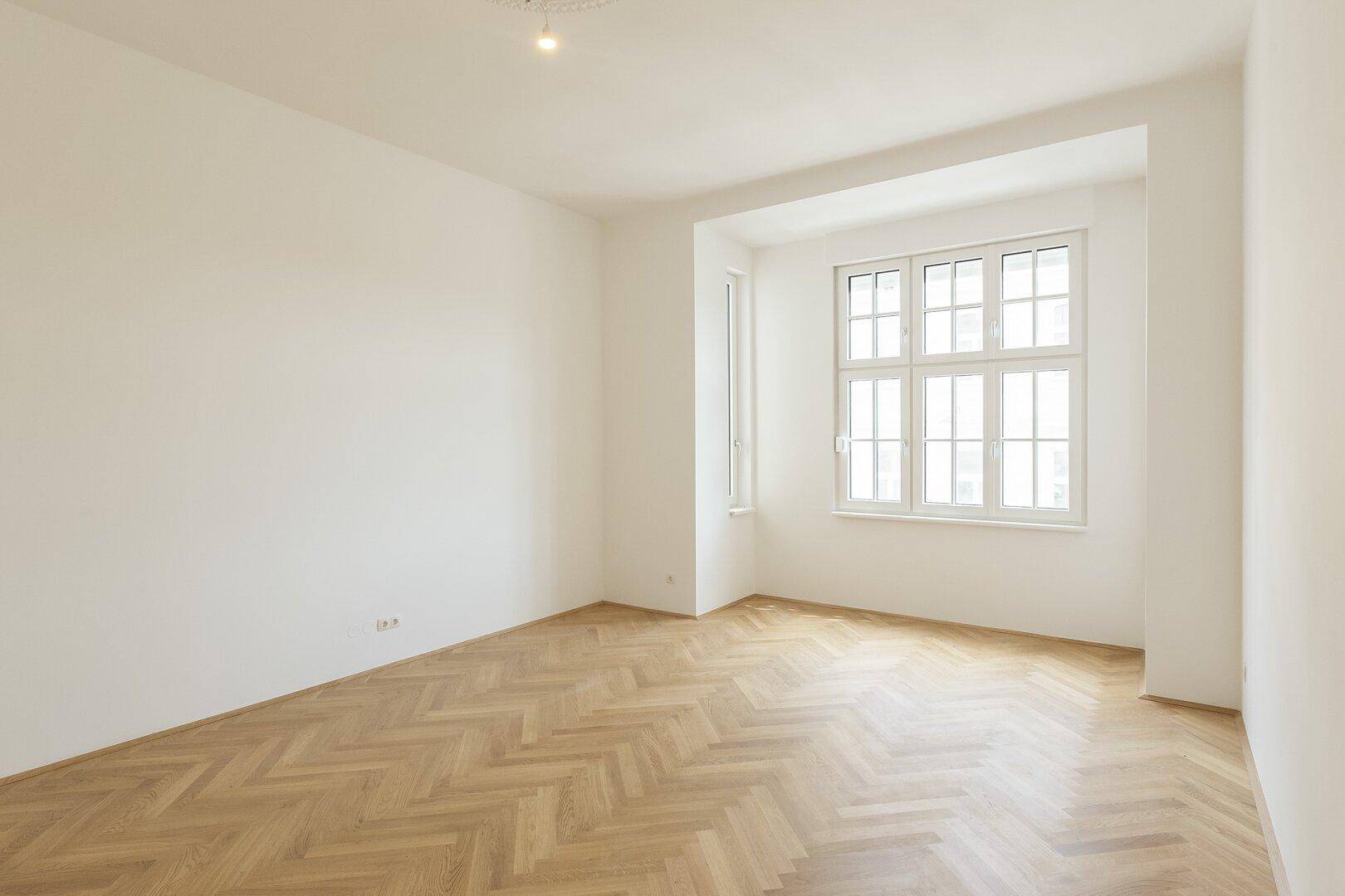Leeres Zimmer mit hellem Fischgrätparkett, weißen Wänden und einem großen Fenster in einer tiefen Fensternische.