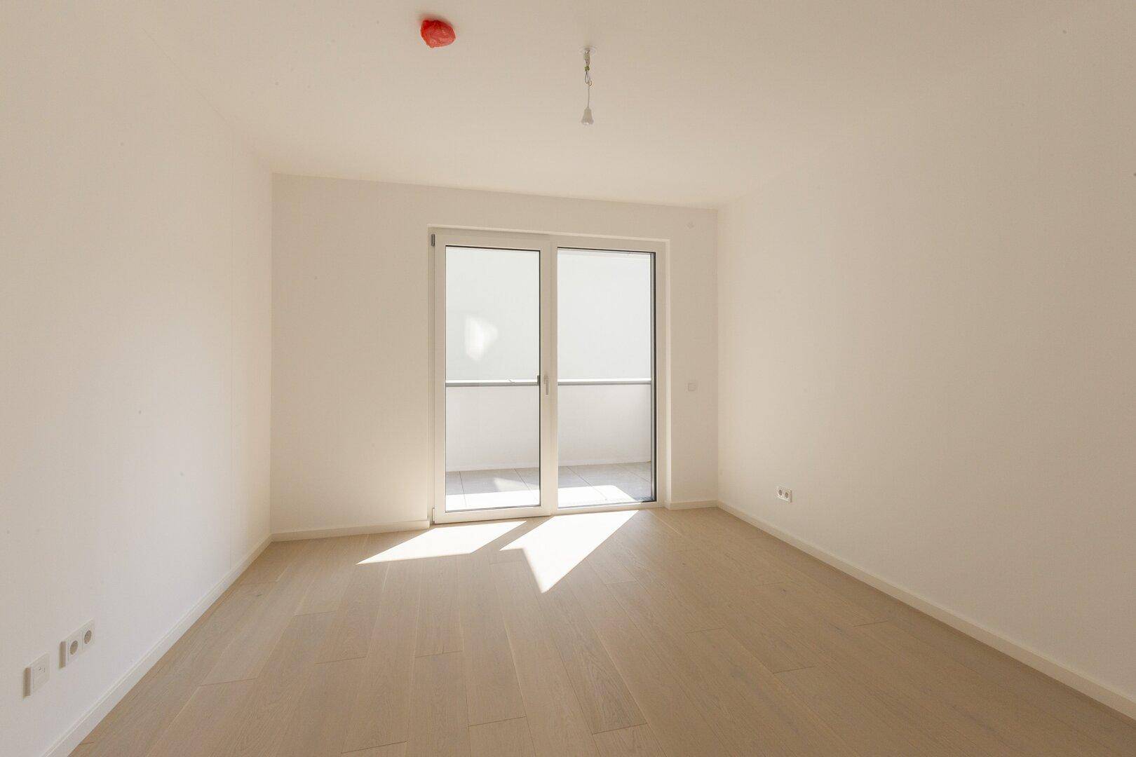 Helles, leeres Zimmer mit Holzboden und Zugang zum Balkon.