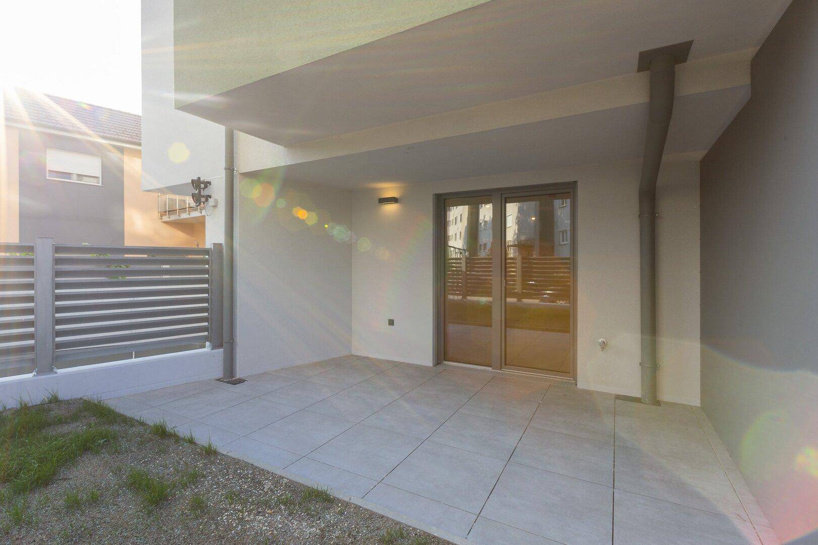 Sonnige, geflieste Terrasse mit Zugang zum Gebäude und moderner Außenbeleuchtung.