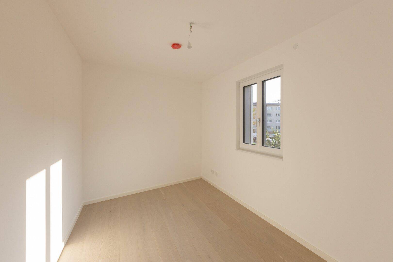 Helles, unmöbliertes Schlafzimmer mit Holzboden und einem Fenster, das viel Tageslicht hereinlässt.