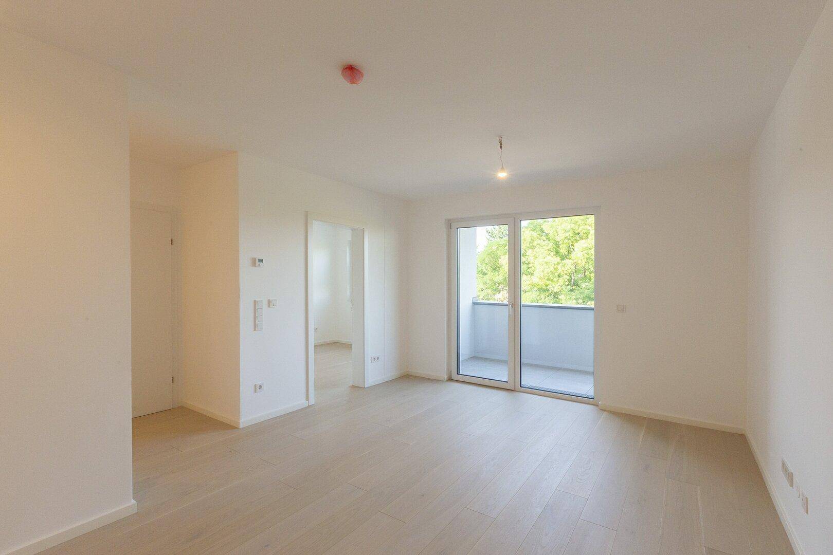 Geräumiger, heller Raum mit Holzboden und großen Fenstern zum Balkon.