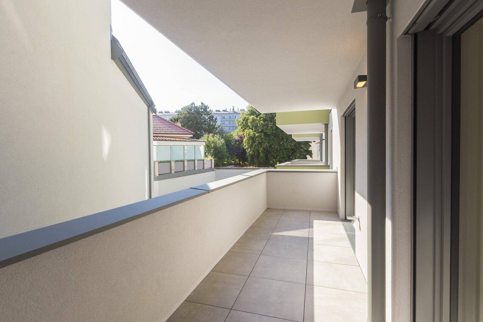 Langer, gefliester Balkon mit Blick auf die umliegenden Gebäude und etwas Grün.
