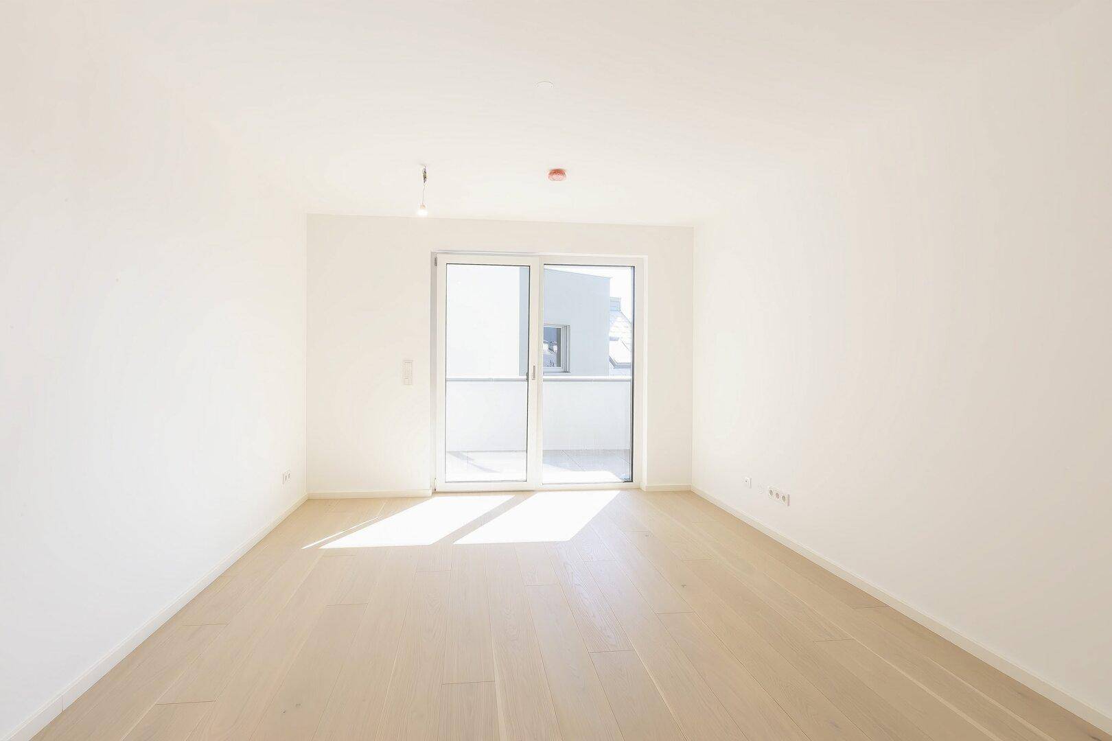Lichtdurchfluteter Raum mit hellem Holzfußboden und Zugang zu einem Balkon.