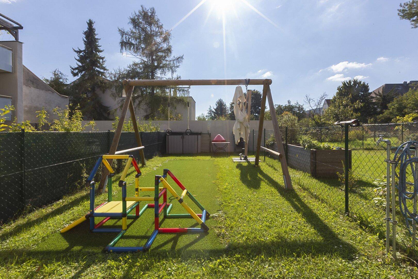 Sonnige Spielplatzfläche mit Schaukel und bunter Kletterstruktur auf Kunstrasen, umgeben von einem Zaun.