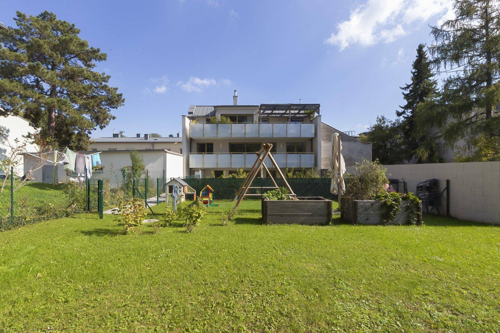Grüner Gartenbereich mit Schaukel, Hochbeeten und einem Mehrfamilienhaus mit Balkonen im Hintergrund unter blauem Himmel.