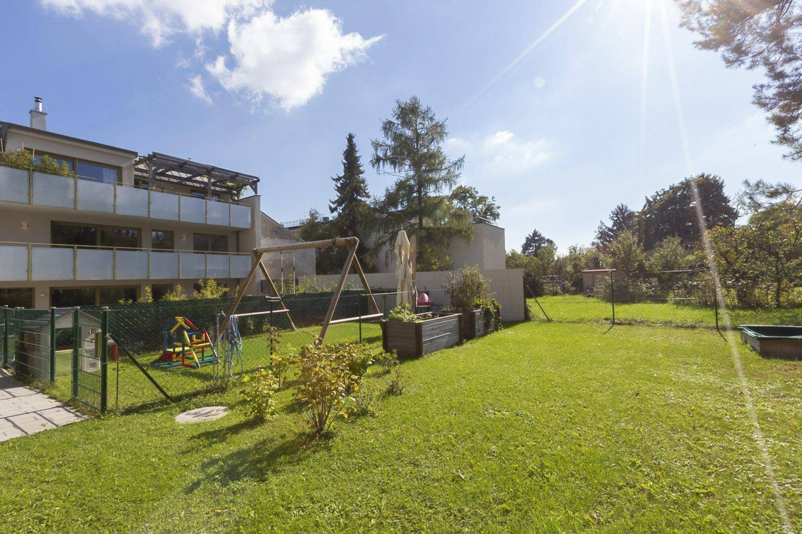 Weitläufige Rasenfläche mit einer Schaukel und einem Wohngebäude mit Balkonen im Hintergrund an einem sonnigen Tag.