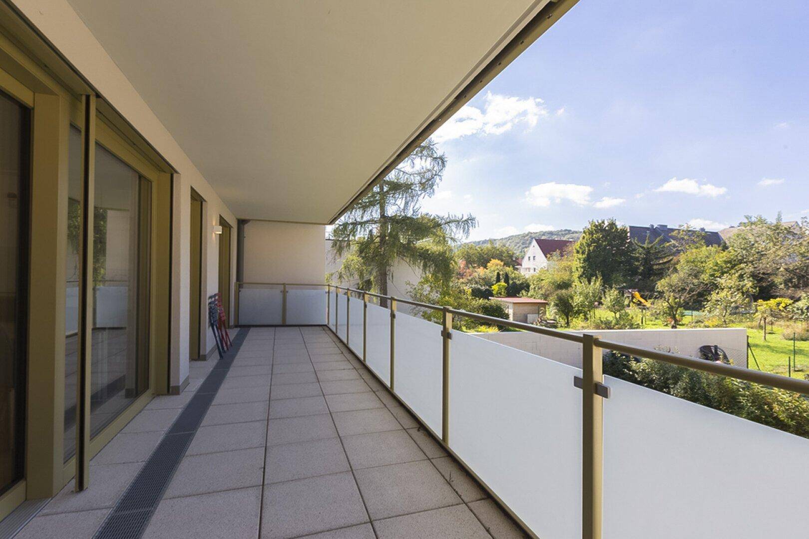 Langer Balkon mit Fliesenboden, Glasgeländer und weitem Blick ins Grüne unter blauem Himmel.