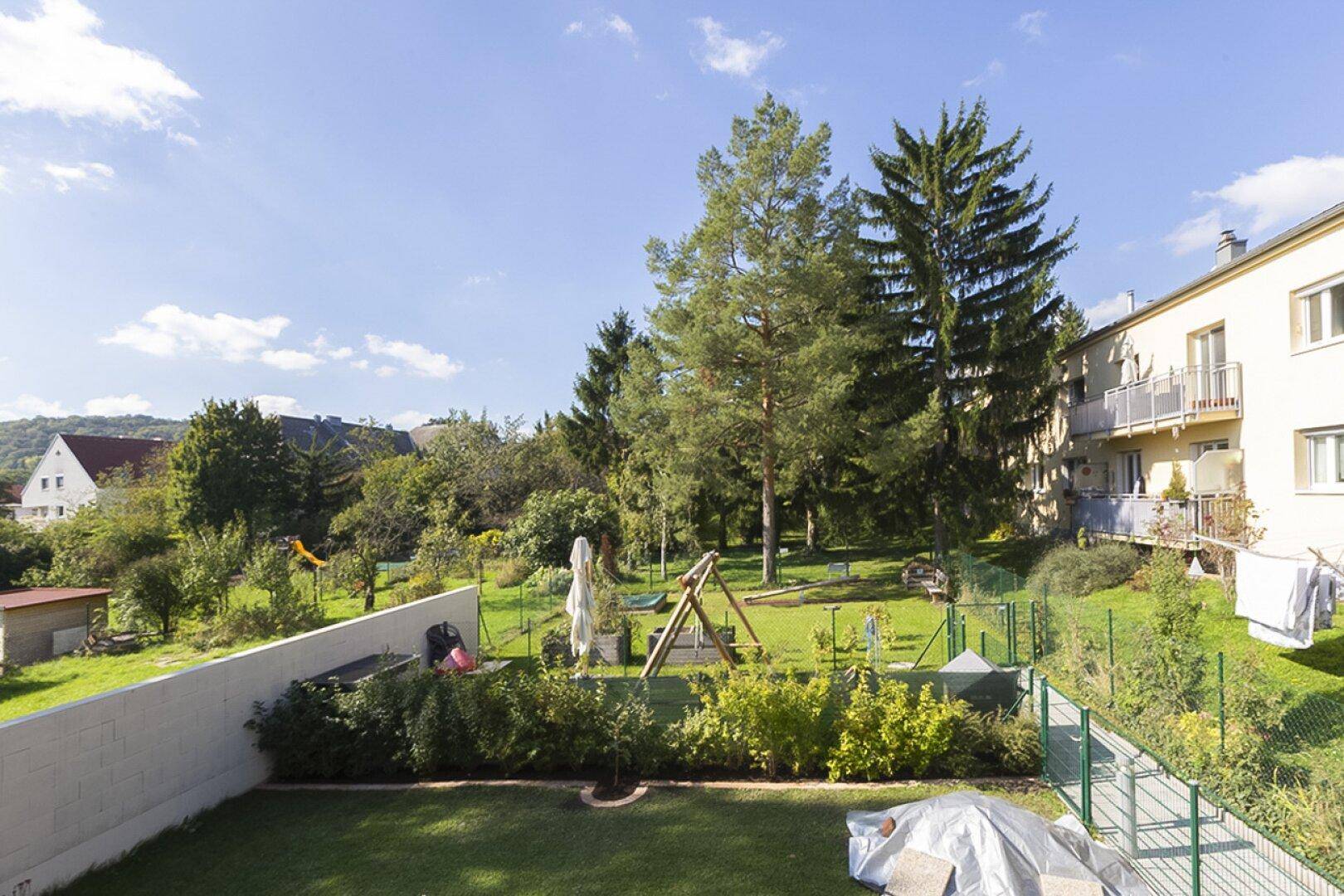 Großer Gemeinschaftsgarten mit Rasenfläche, Bäumen und einem Spielplatz, umgeben von Wohngebäuden unter blauem Himmel.
