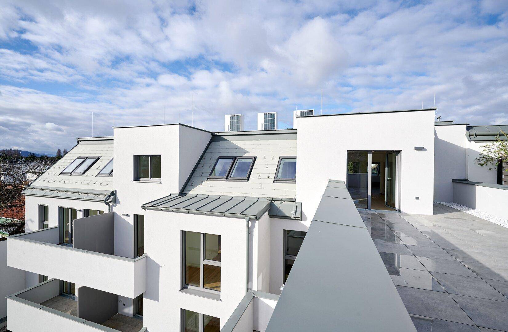 Moderne Dachterrasse mit Blick auf die weiße Fassade des Neubaus und die umliegende Landschaft.