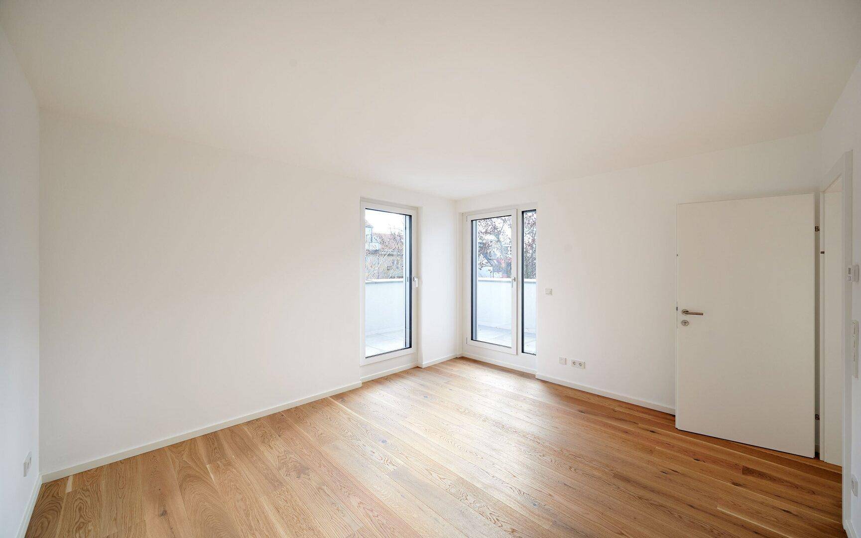 Helles Schlafzimmer mit Holzboden und zwei Fenstern, die Zugang zu einem französischen Balkon bieten.