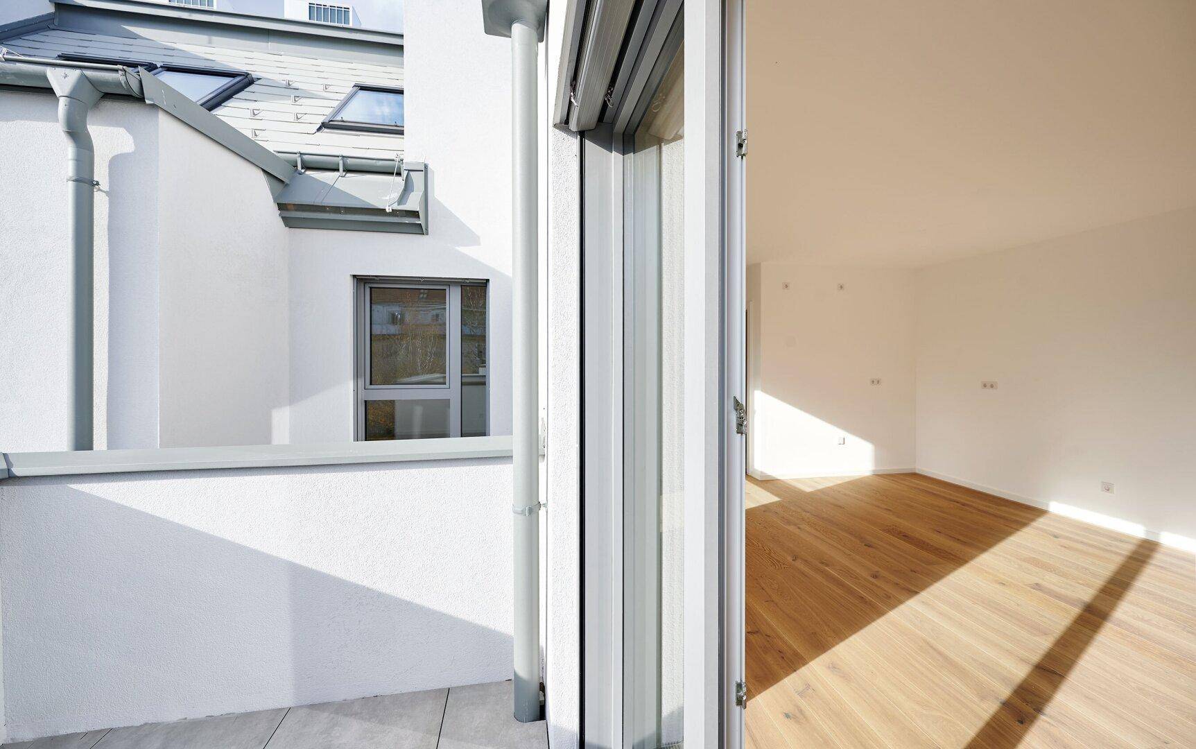 Blick aus dem Wohnzimmer auf den Balkon mit Fliesenboden und die umliegende Architektur.