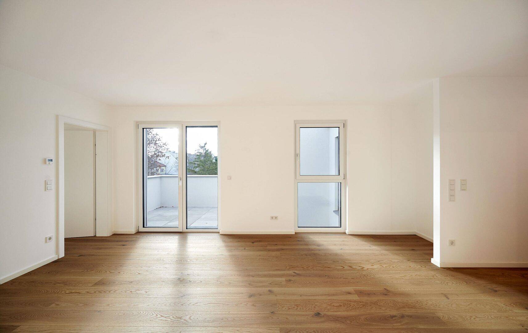 Geräumiges, helles Wohnzimmer mit Holzboden und großen Fenstern, die viel Tageslicht hereinlassen.