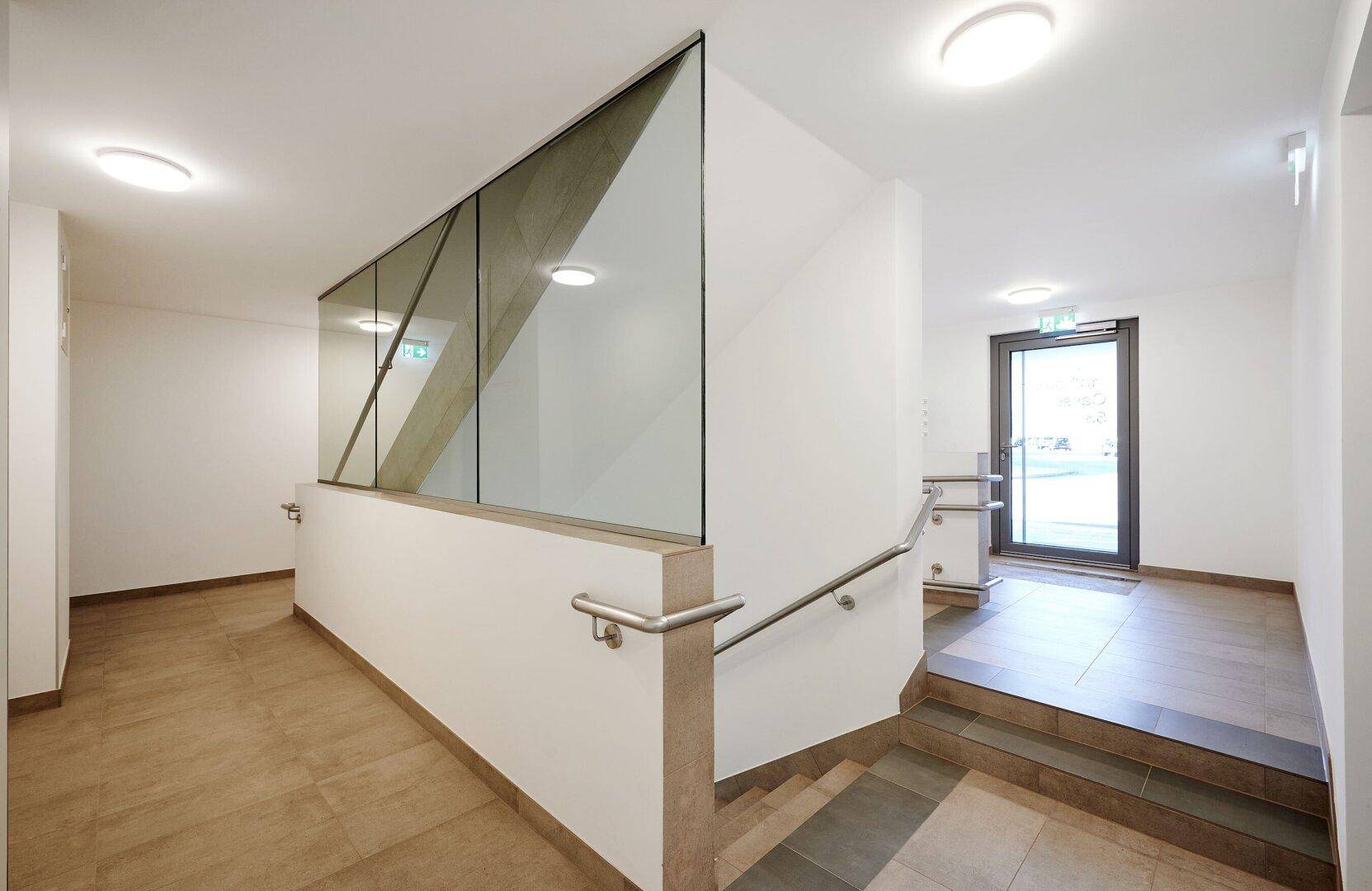 Heller Treppenaufgang mit modernem Glasgeländer und Fliesenboden im Eingangsbereich.