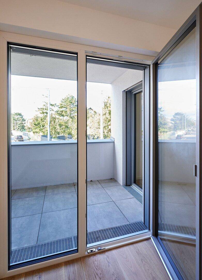 Blick vom Innenraum auf den Balkon mit Fliesenboden und grüner Umgebung.