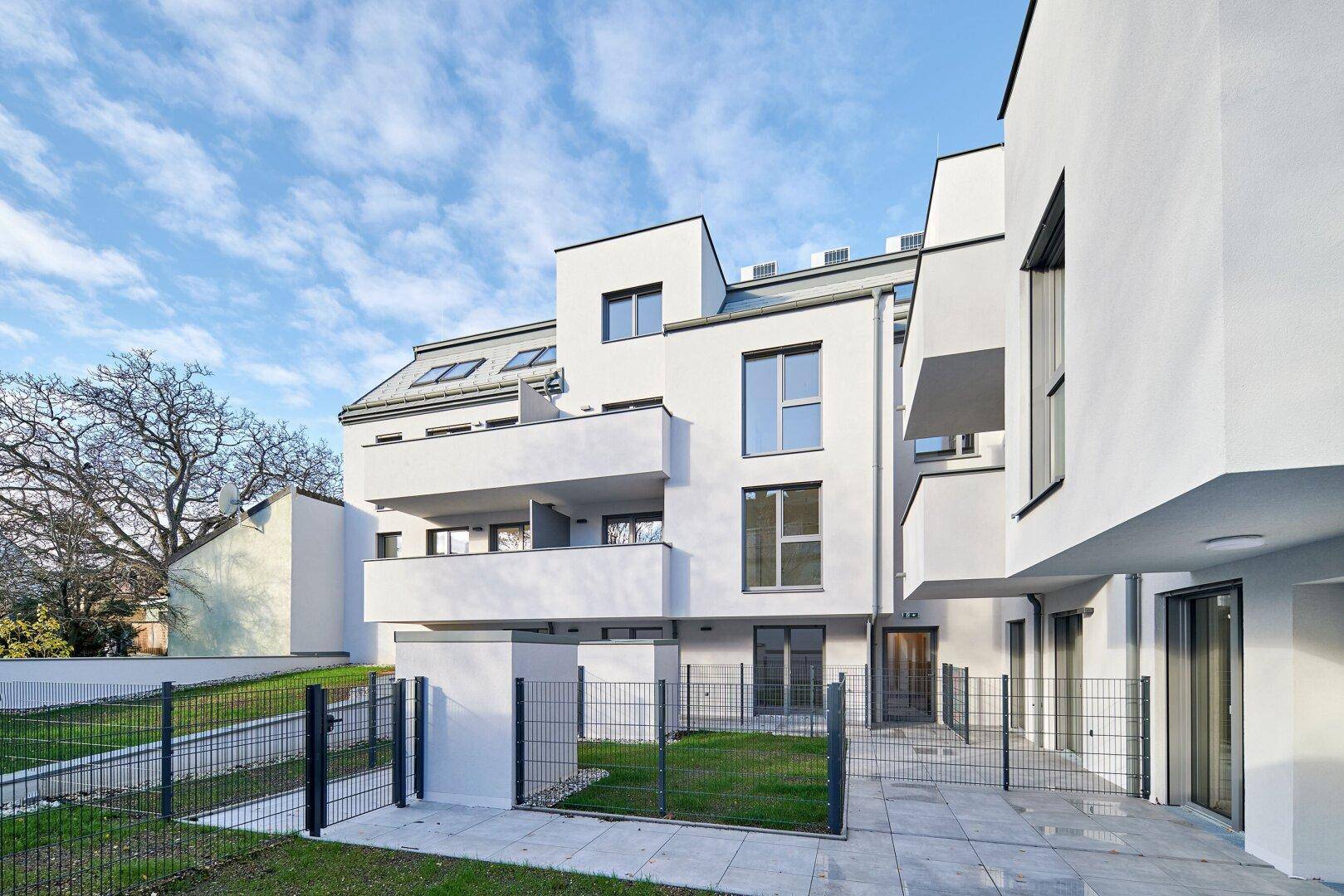 Moderne weiße Wohnanlage mit Balkonen und kleinen Grünflächen unter blauem Himmel.