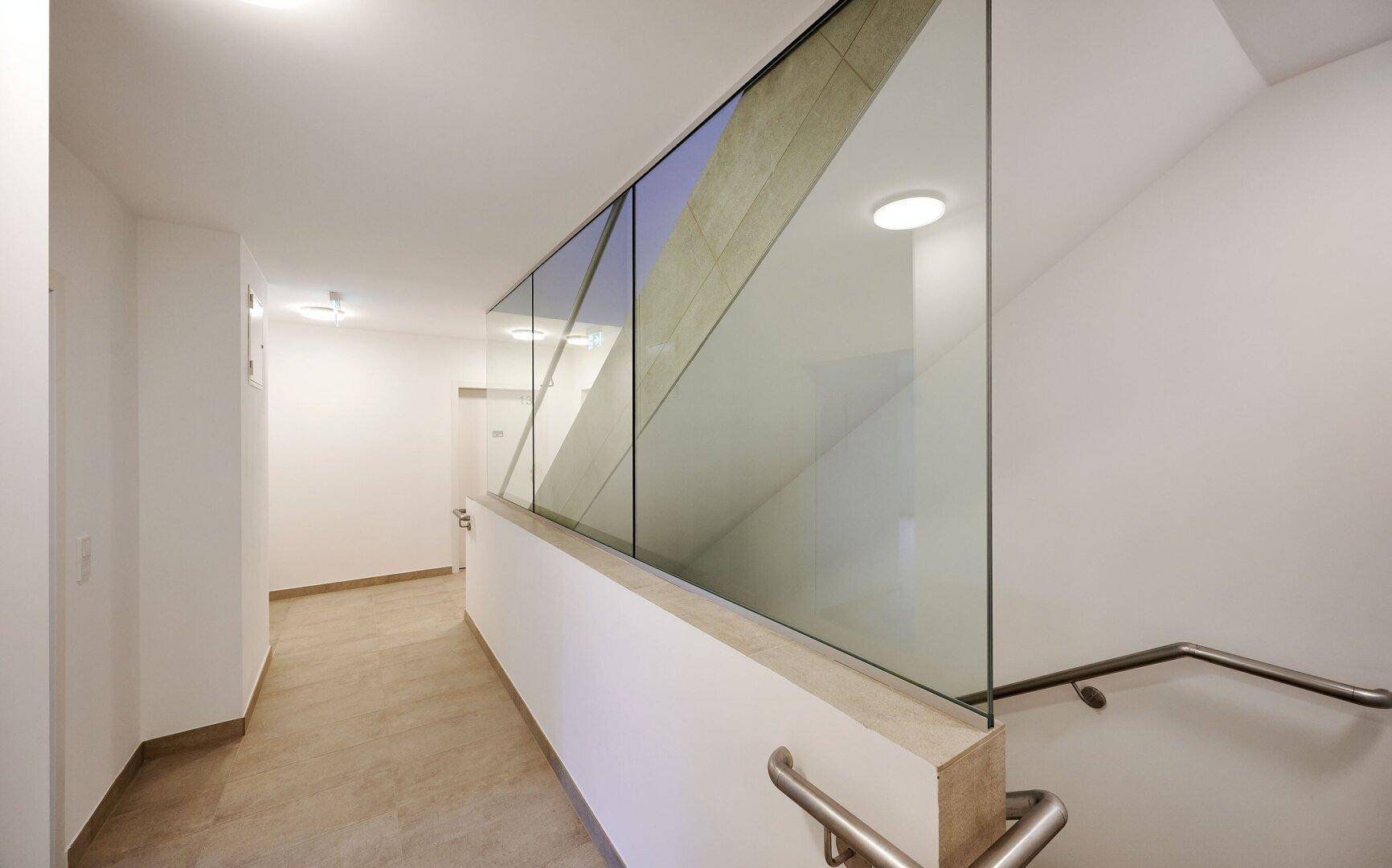 Heller Gemeinschaftsflur mit modernem Glasgeländer und Fliesenboden, der zu den Wohnungen führt.