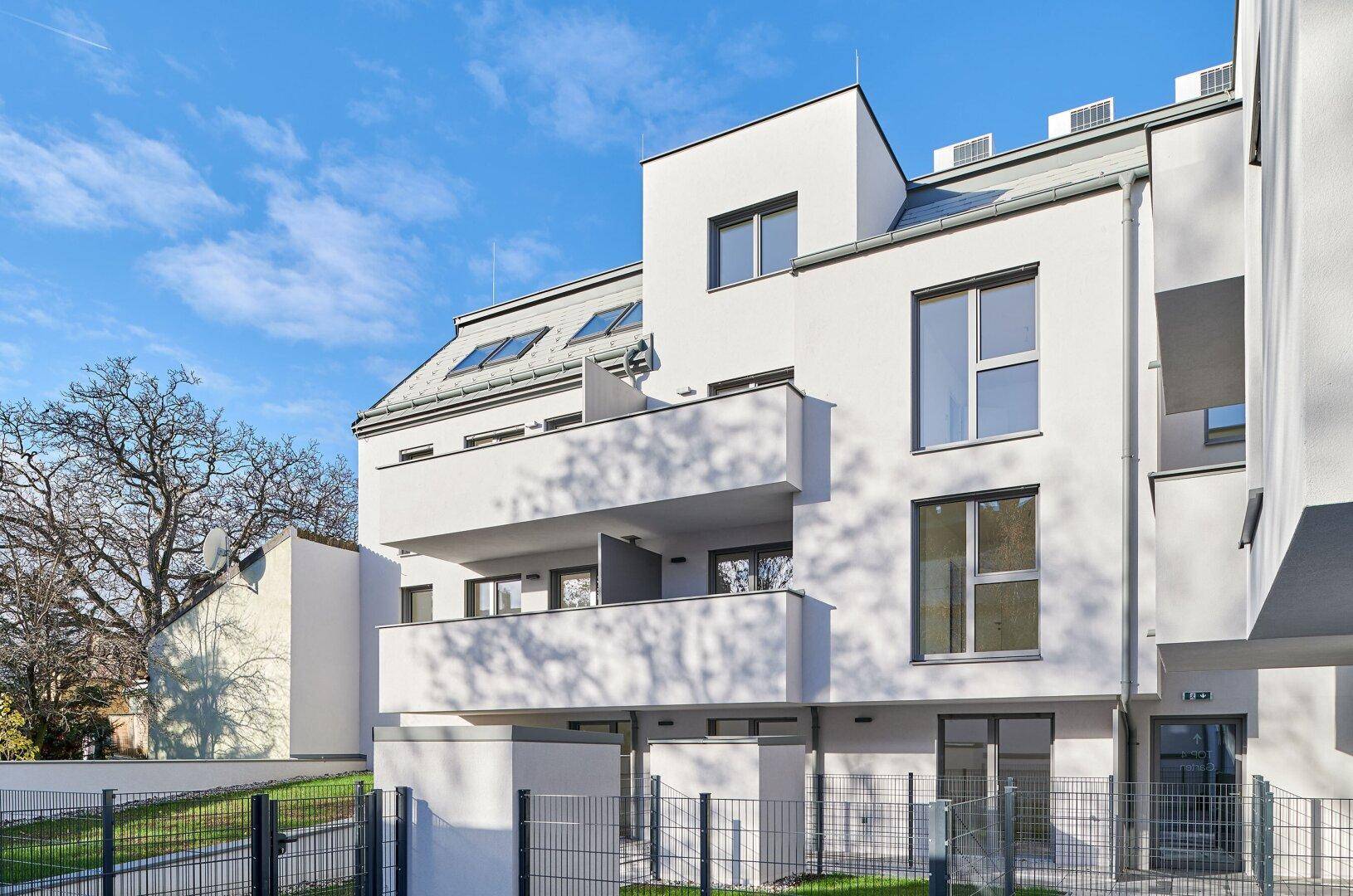 Seitenansicht eines modernen Wohngebäudes mit Balkonen, weißer Fassade und einem kleinen Gartenbereich.