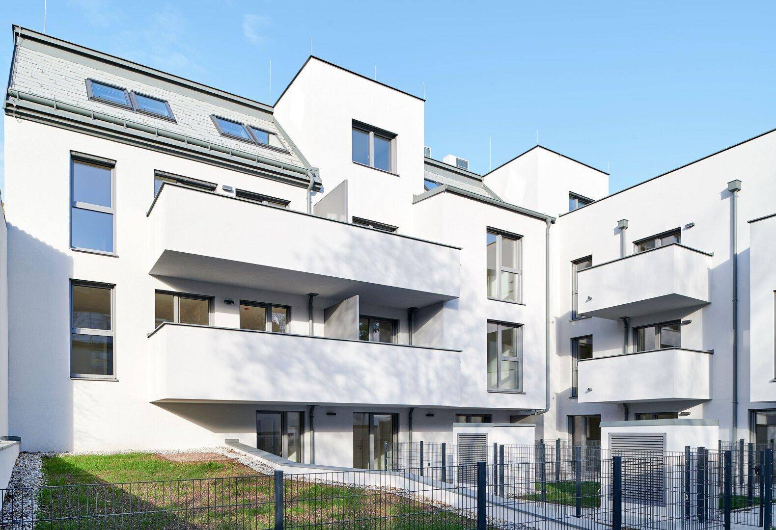 Rückansicht eines modernen Wohnkomplexes mit Balkonen, weißer Fassade und begrünten Außenflächen.