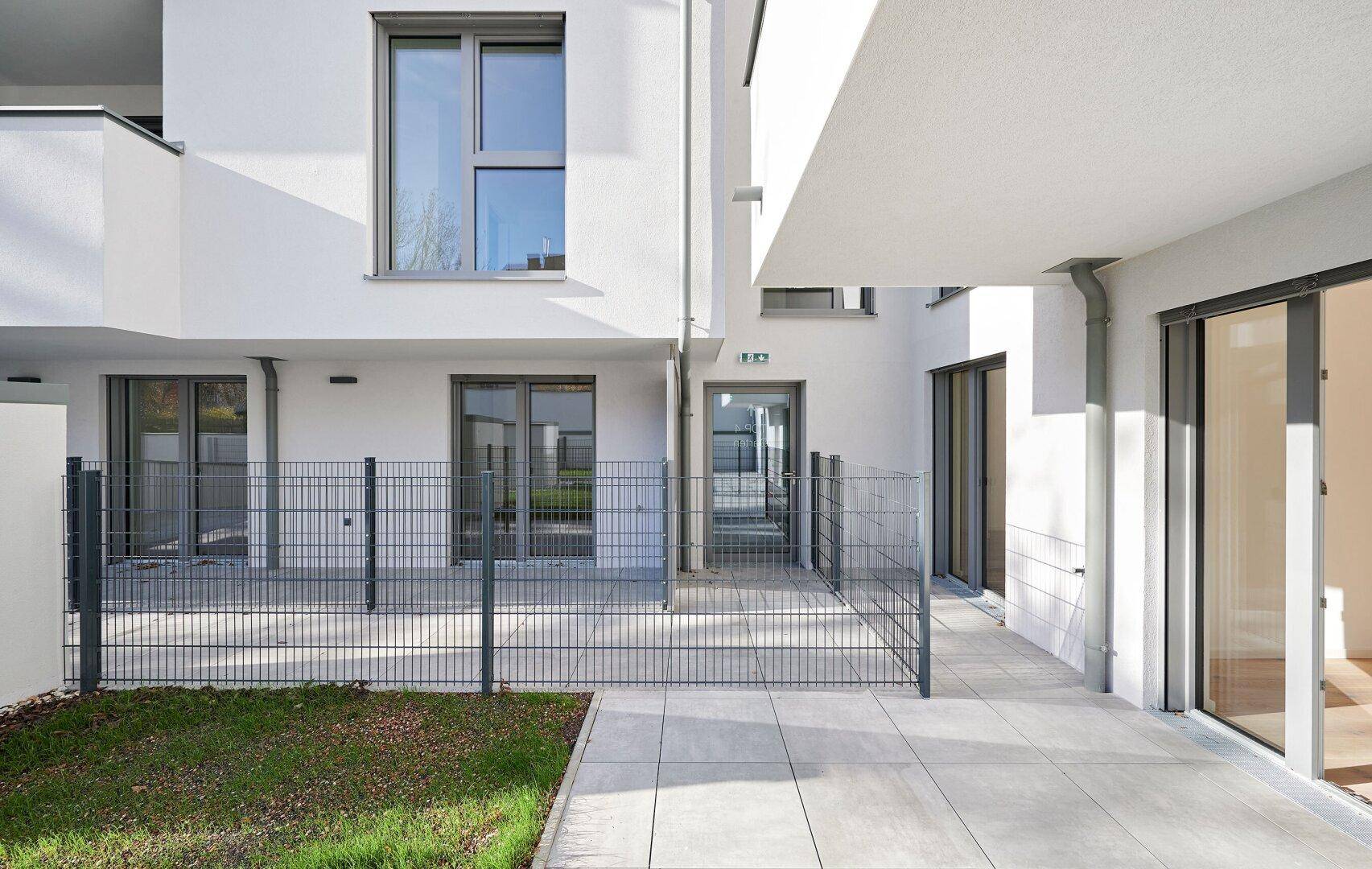 Moderne Terrasse mit Steinplatten und kleinem Rasenbereich, umzäunt und zugänglich vom Gebäude.