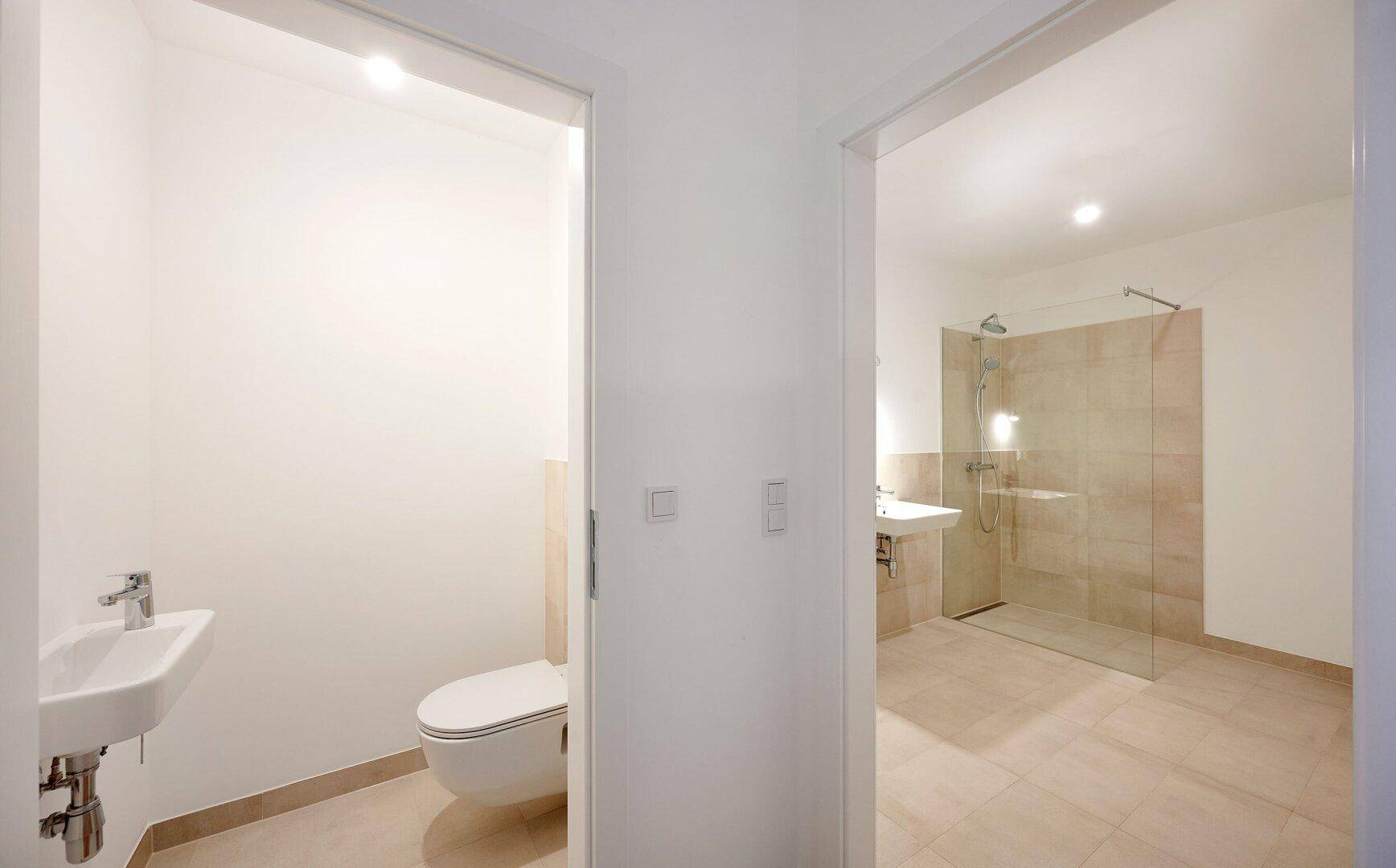 Blick vom Flur in ein separates Gäste-WC und ein modernes Badezimmer mit Dusche.