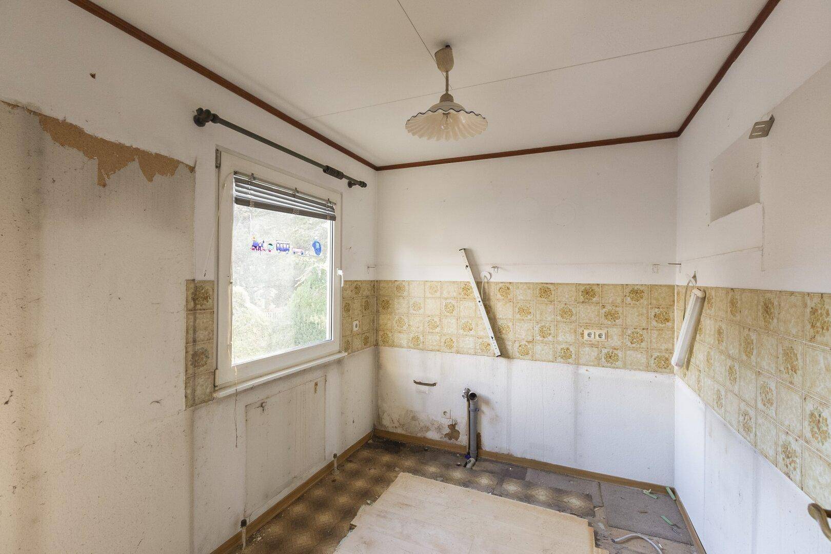 Leere Küche mit Fenster, alten Fliesen und sichtbaren Anschlüssen, bereit für eine Renovierung.