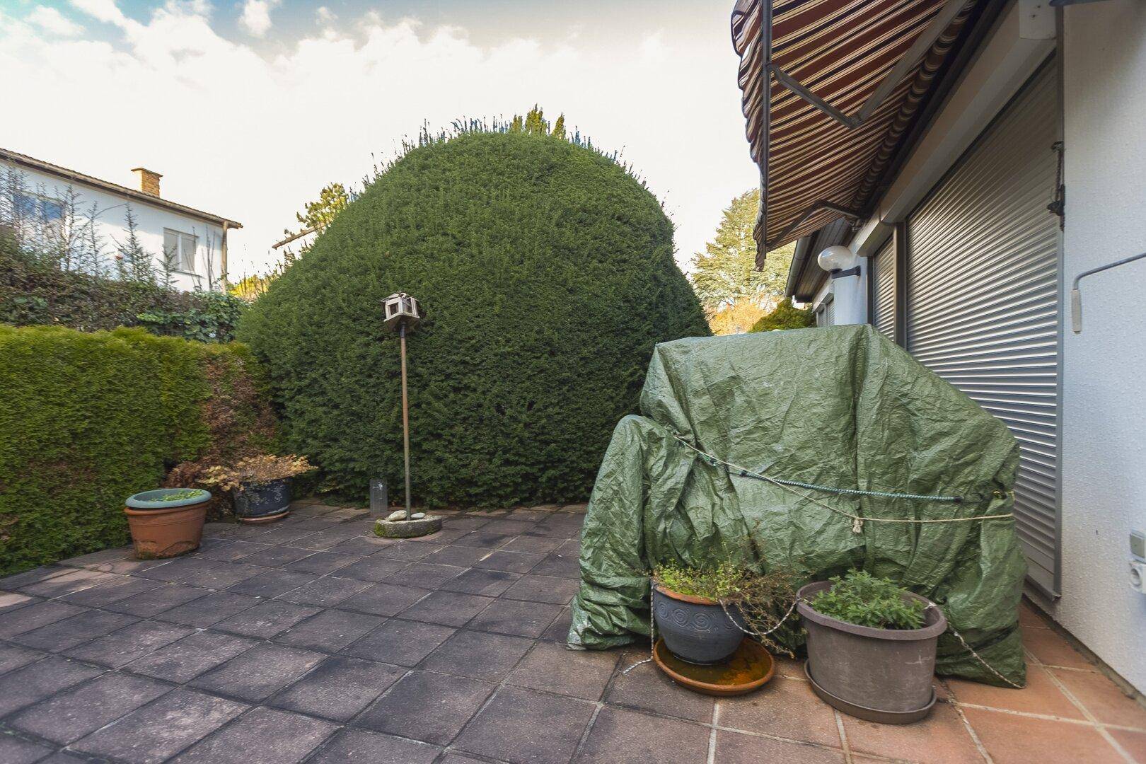 Gepflasterte Terrasse mit Topfpflanzen und einer abgedeckten Sitzgelegenheit, umgeben von hohen Hecken.