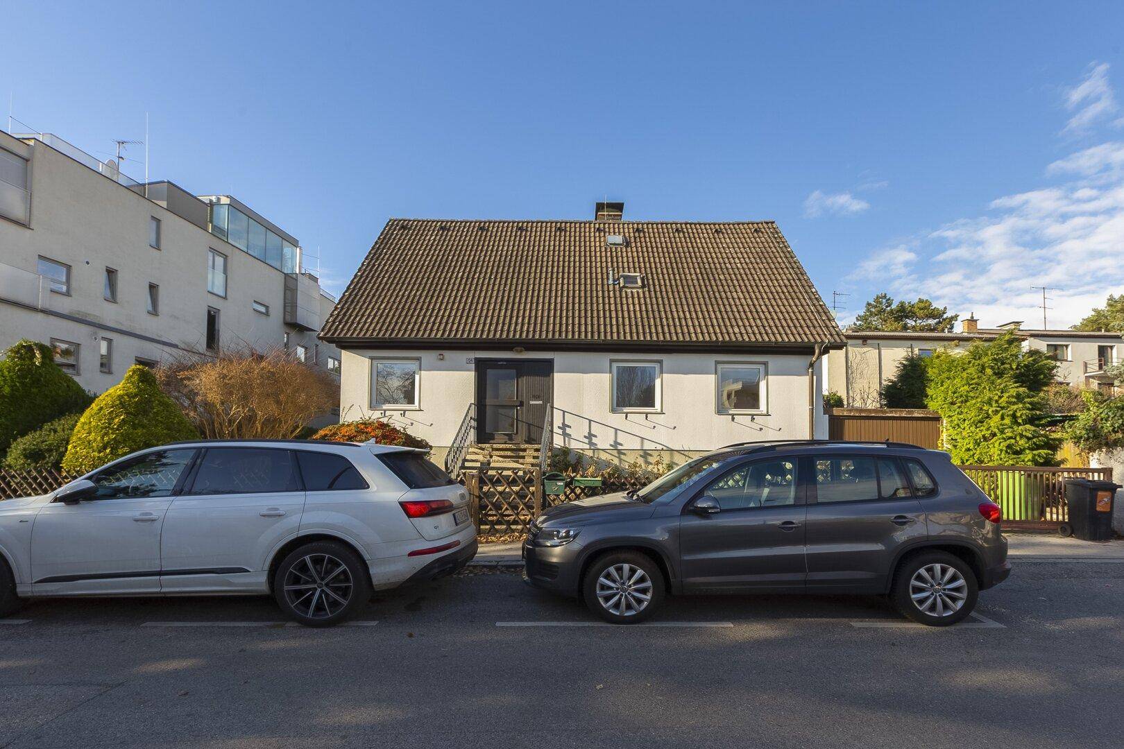Einfamilienhaus von der Straße aus gesehen, mit parkenden Autos davor und umliegenden Gebäuden.