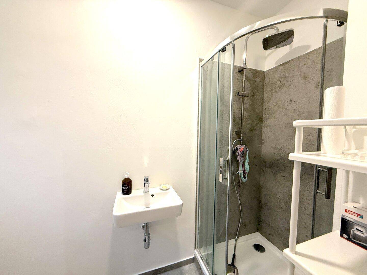 Modernes Badezimmer mit Duschkabine, kleinem Waschbecken und grauen Fliesen.