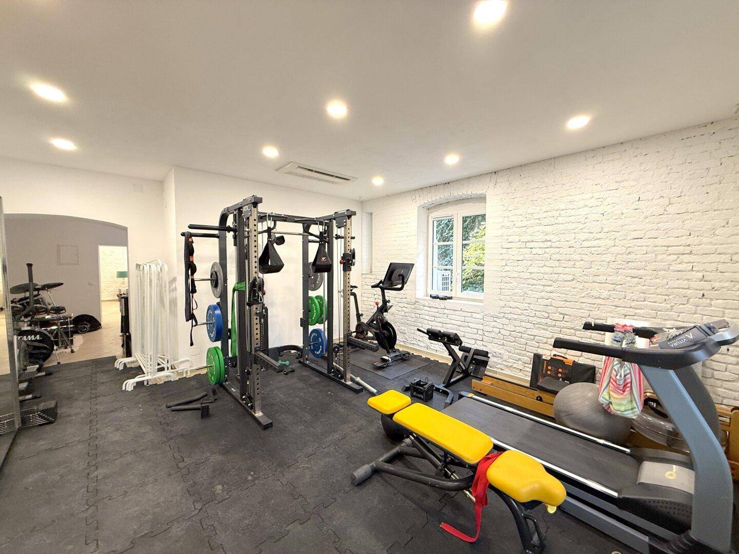 Fitnessraum mit verschiedenen Trainingsgeräten, Gummiboden und weiß gestrichenen Ziegelwänden.