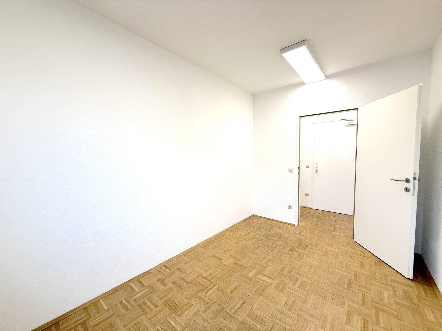 Leerer Büroraum mit Parkettboden und weißen Wänden, Blick durch eine offene Tür in einen weiteren Raum.