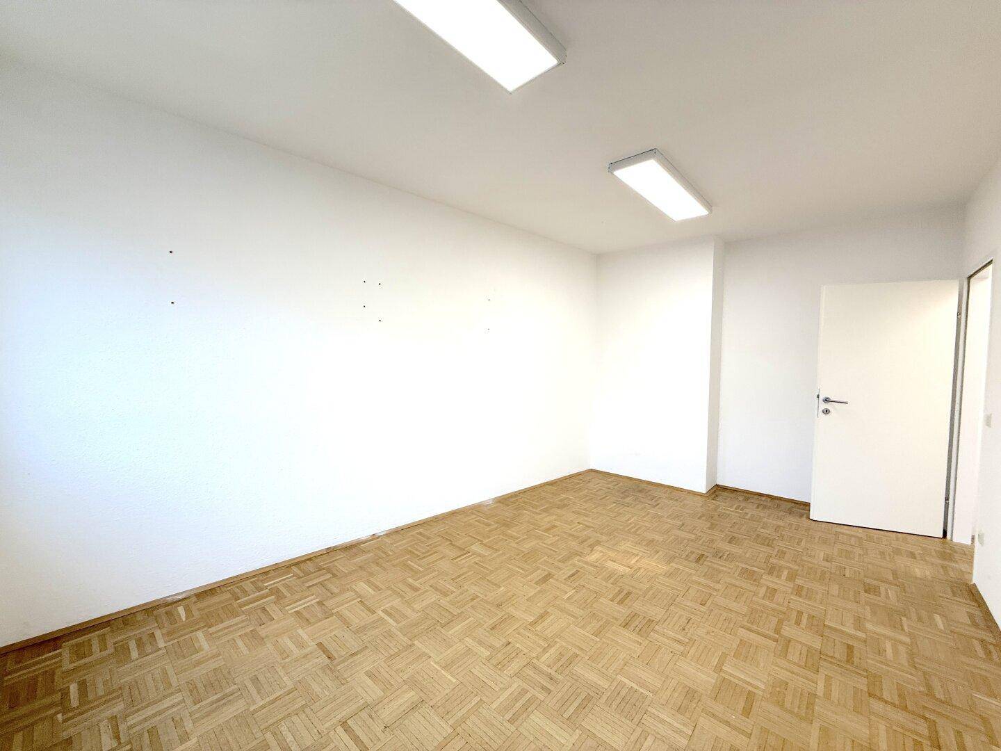 Leerer, heller Büroraum mit Parkettboden, weißen Wänden und einer Tür.