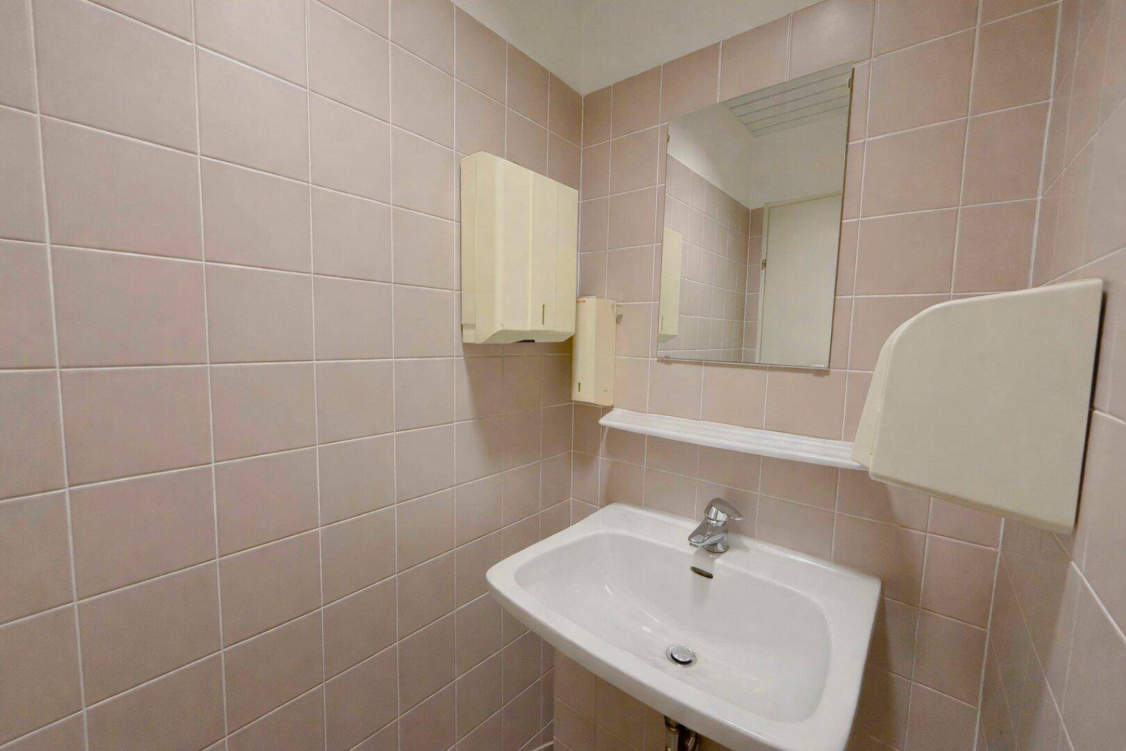 Kleiner Toilettenraum mit Waschbecken, Spiegel und hellbraunen Fliesen an den Wänden.