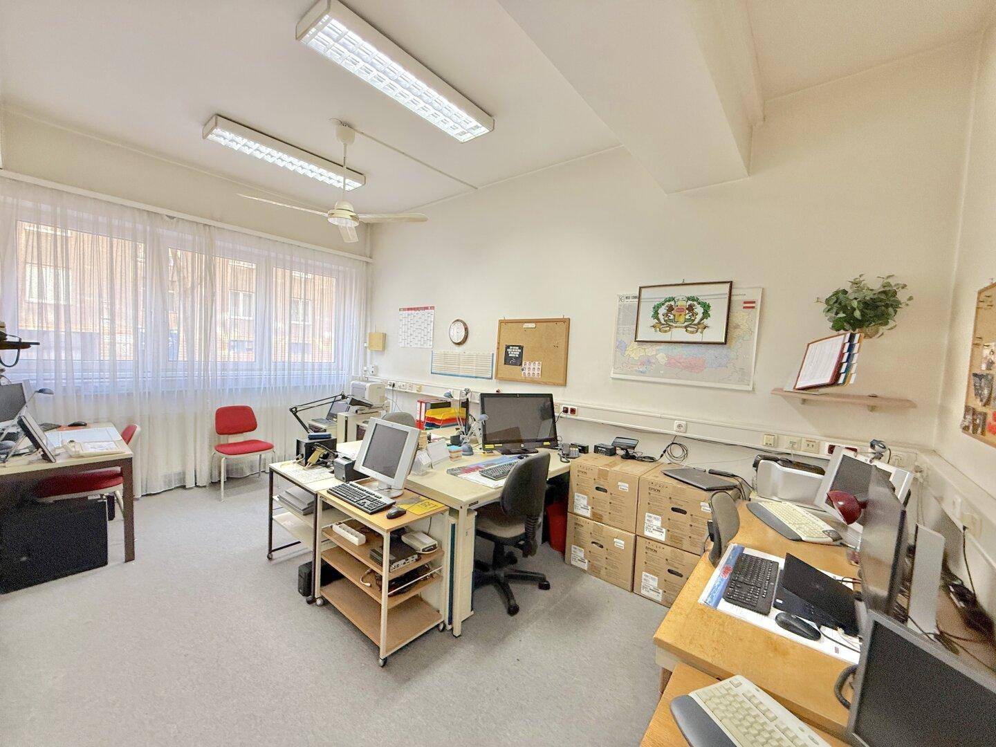 Helles Großraumbüro mit mehreren Arbeitsplätzen, Teppichboden und Deckenventilator für angenehmes Klima.
