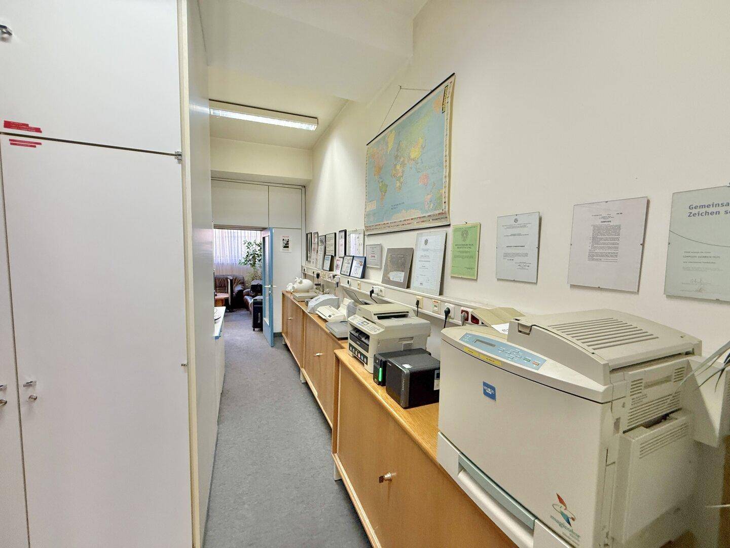 Langer Flur mit Aktenschränken, Druckern und einer Weltkarte an der Wand, führt zu weiteren Räumen.