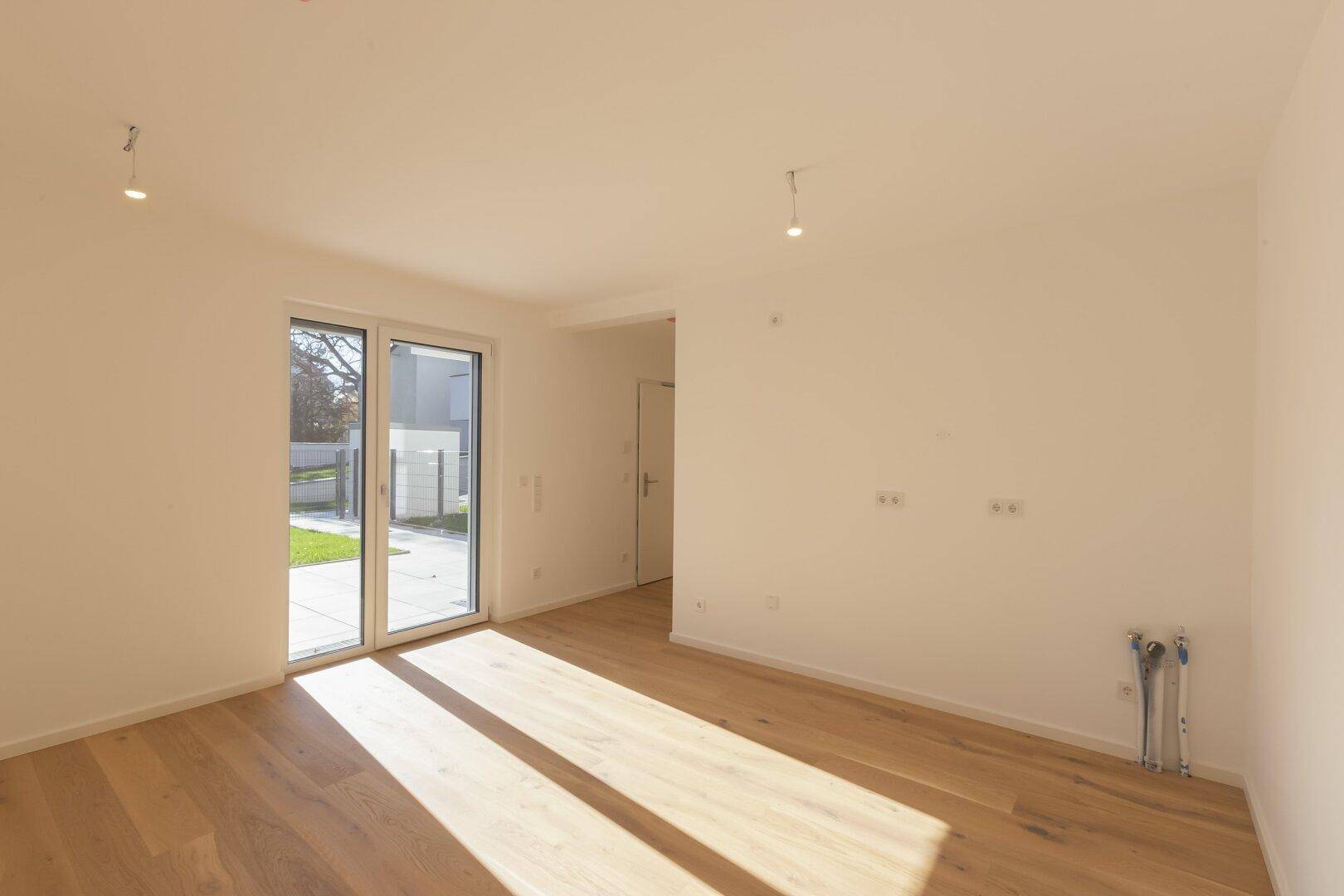 Helles Wohnzimmer mit Holzboden, Zugang zum Garten und vorbereiteten Küchenanschlüssen.