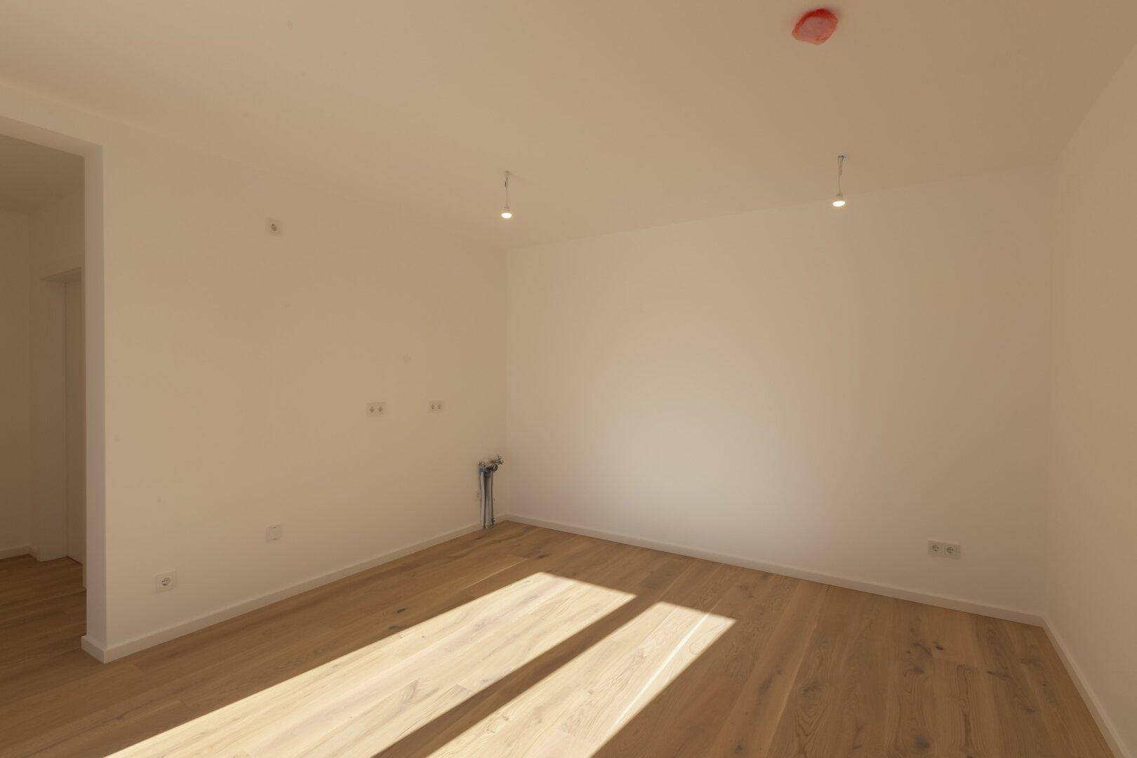 Geräumiges Wohnzimmer mit Holzboden und Anschlüssen für eine offene Küche.