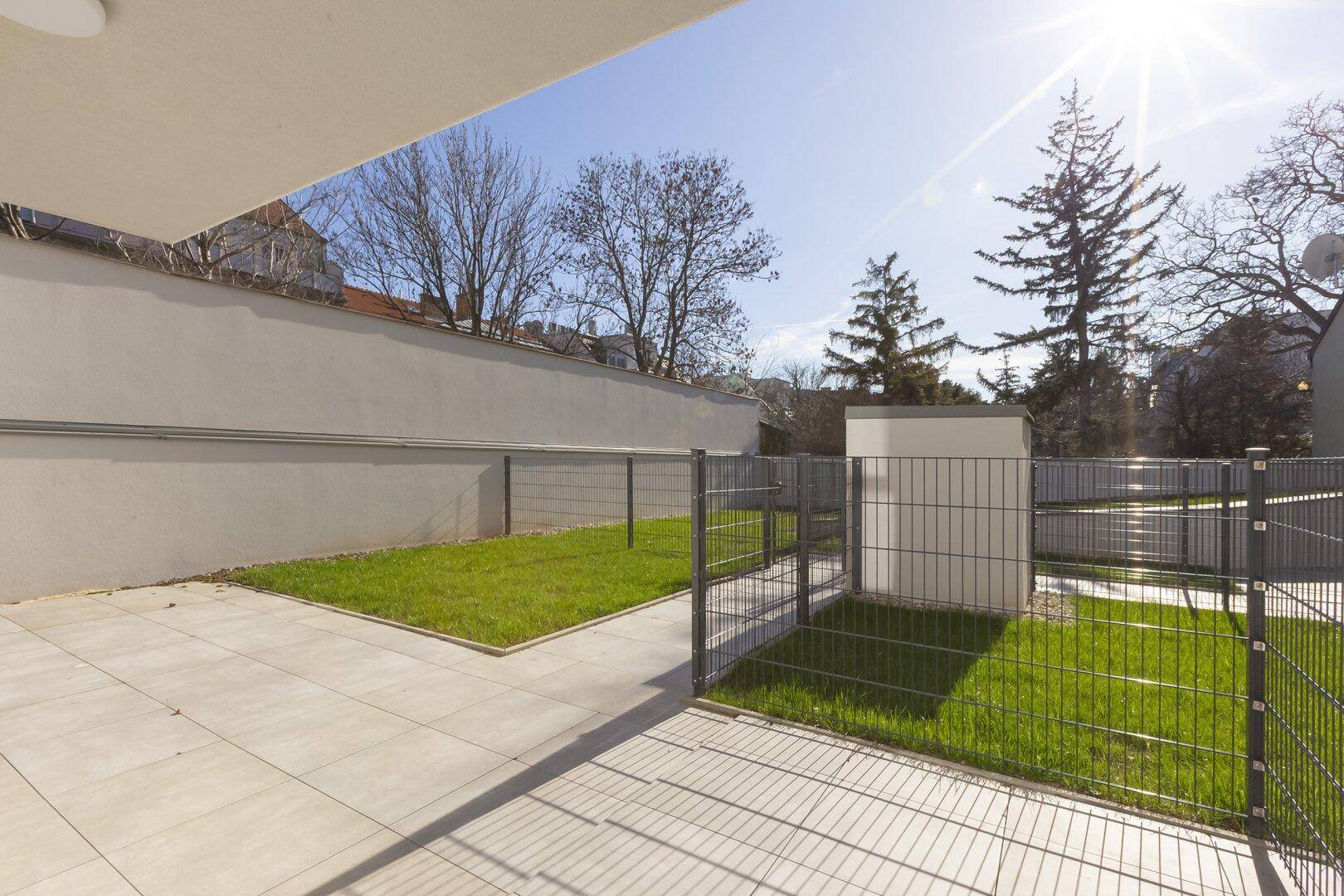 Sonniger Außenbereich mit gepflasterter Terrasse und kleinem Rasenstück, umgeben von einem Zaun.