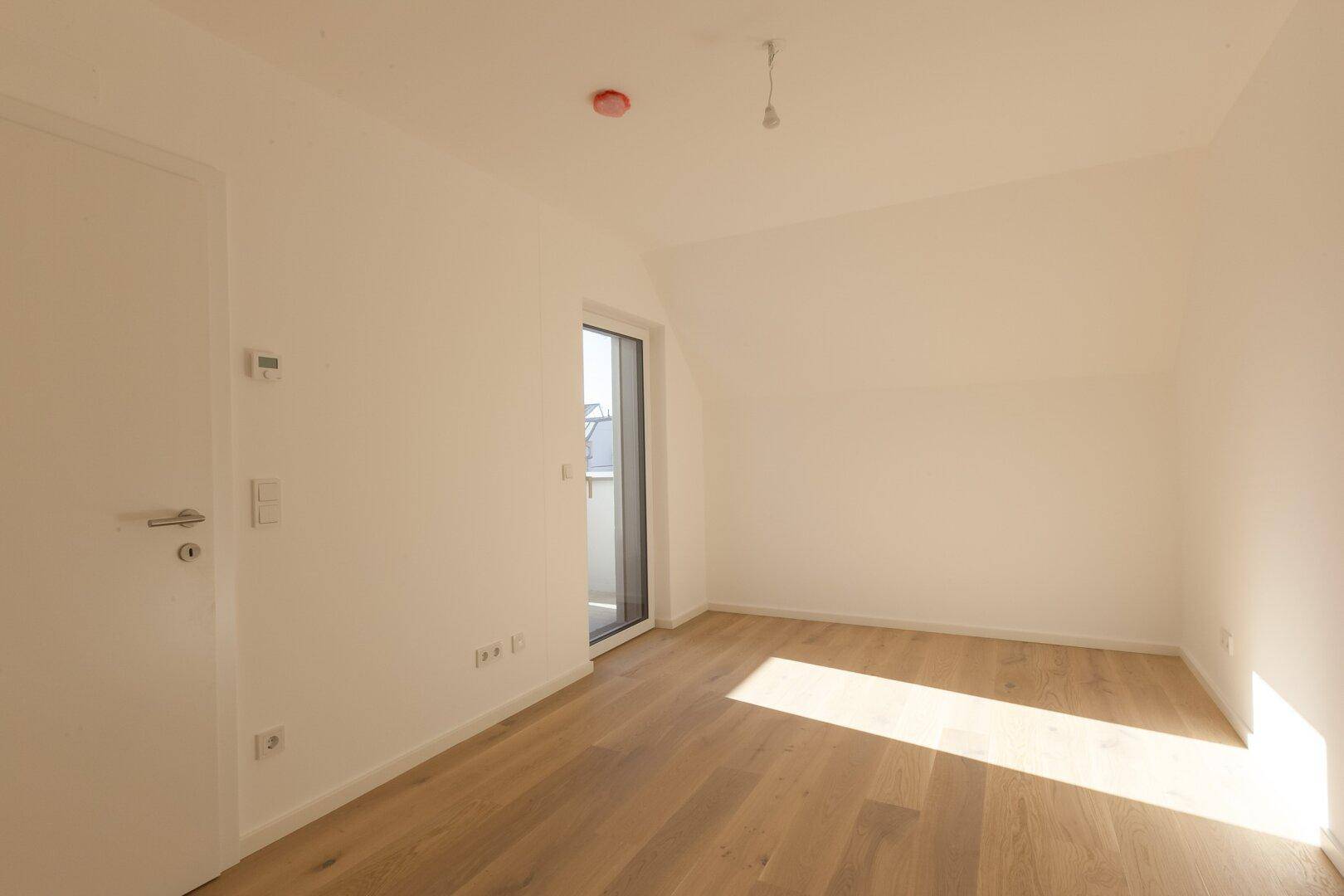 Lichtdurchflutetes Zimmer mit Holzboden und Tür zu einem kleinen Balkon, bietet viel Helligkeit.