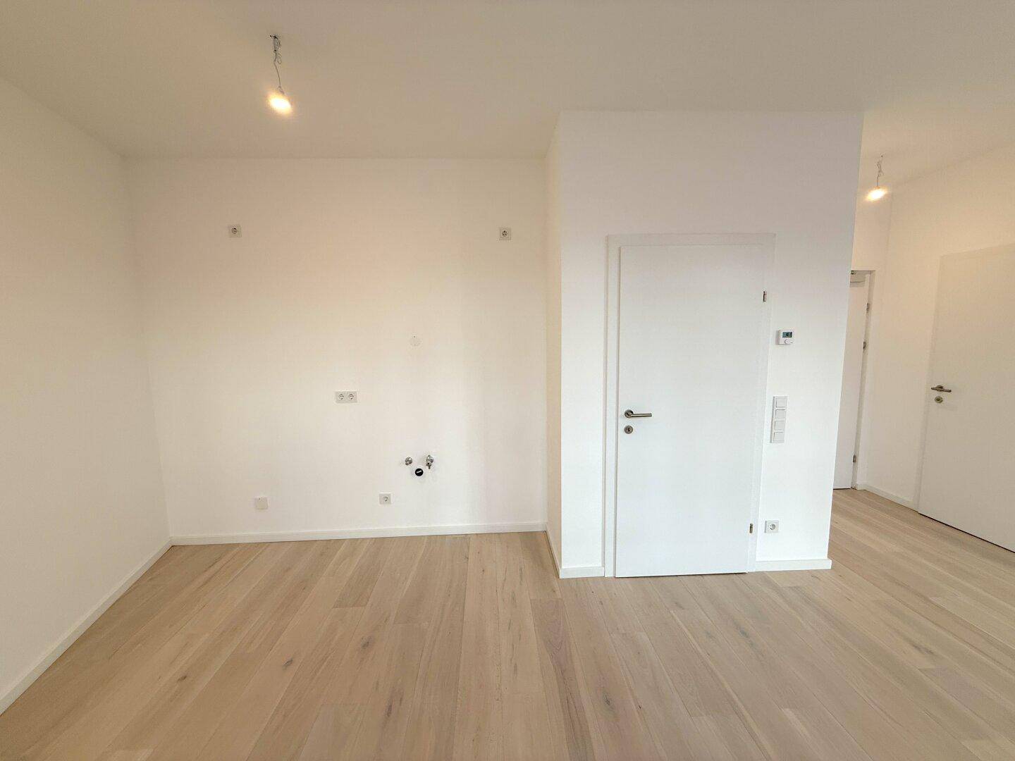 Leerer Küchenbereich mit Anschlüssen für eine Einbauküche und hellem Holzboden.