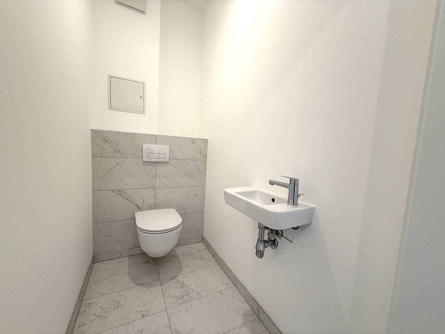 Modernes Gäste-WC mit Wand-WC, kleinem Waschbecken und hellen Fliesen.
